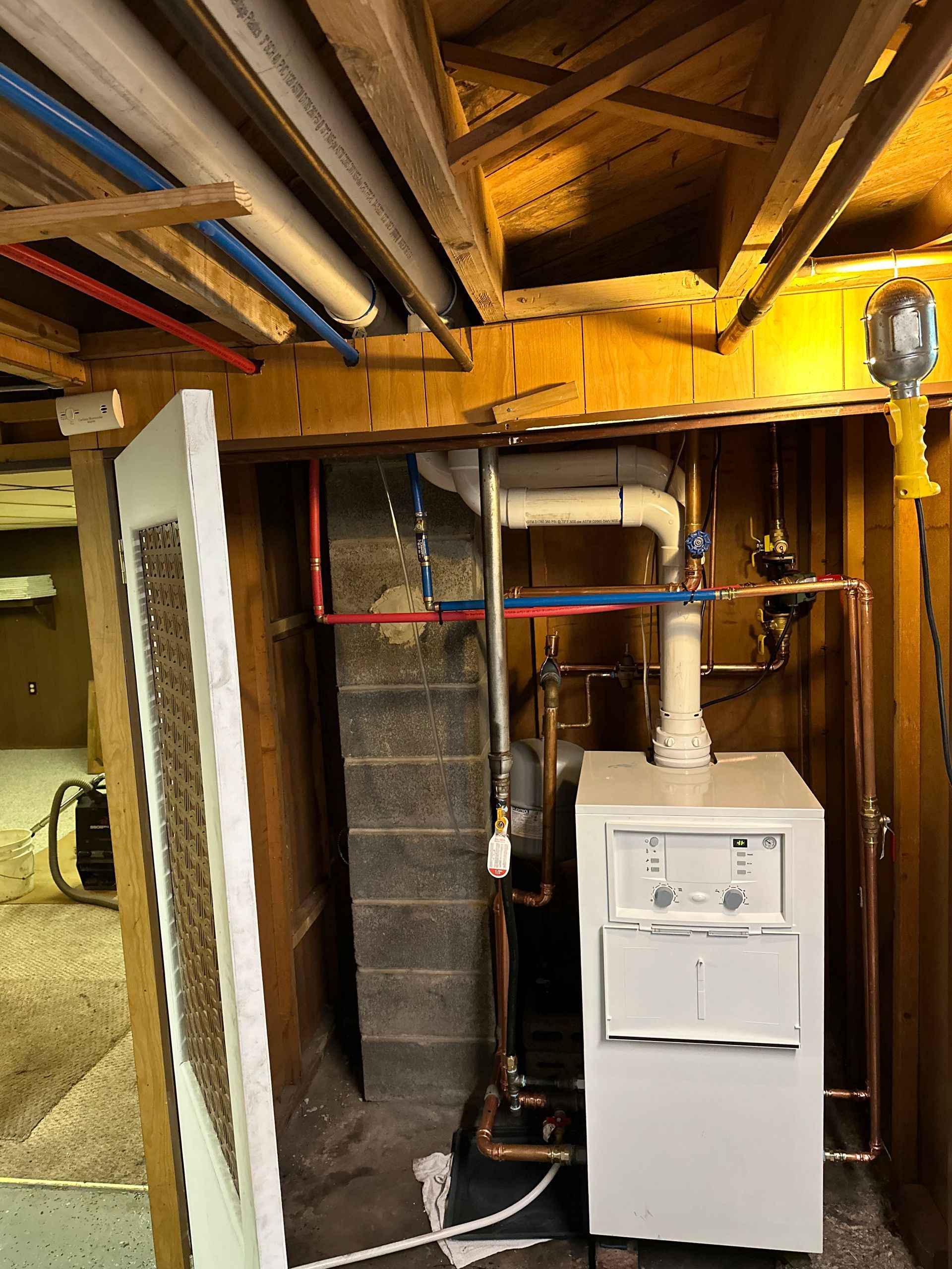 White furnace in a basement with copper pipes and wood beams. Red and blue water lines on the wall.