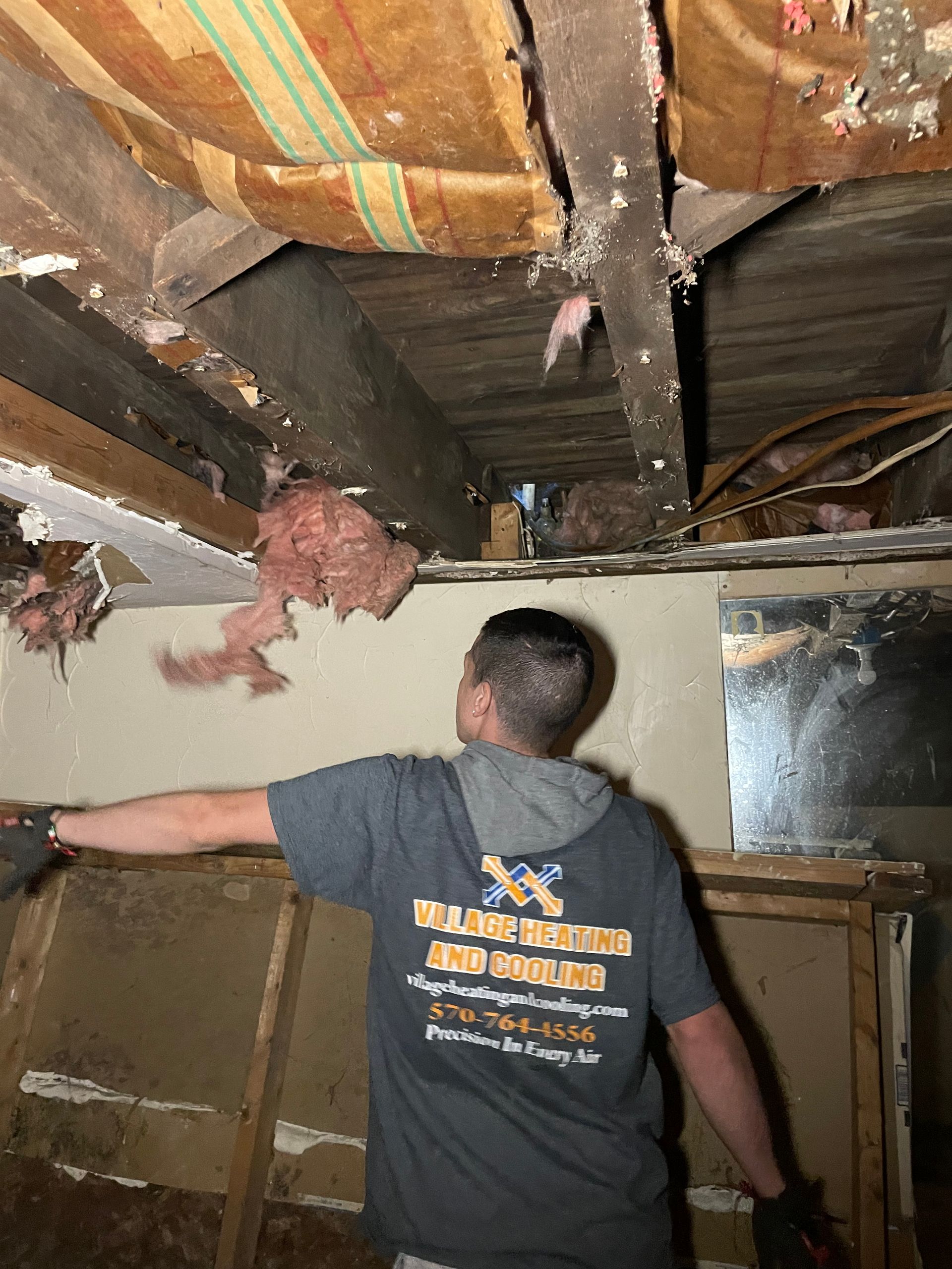 Man in gray shirt reaching up at damaged ceiling, likely in a basement.