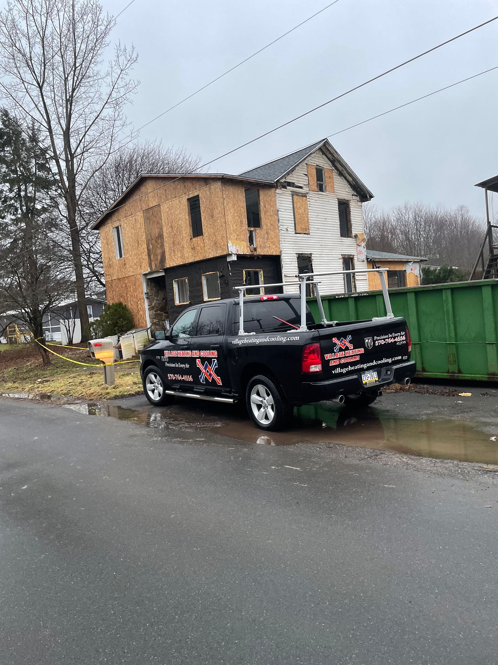Black truck parked on a wet street in front of a dilapidated house. Green dumpster nearby. Overcast sky.