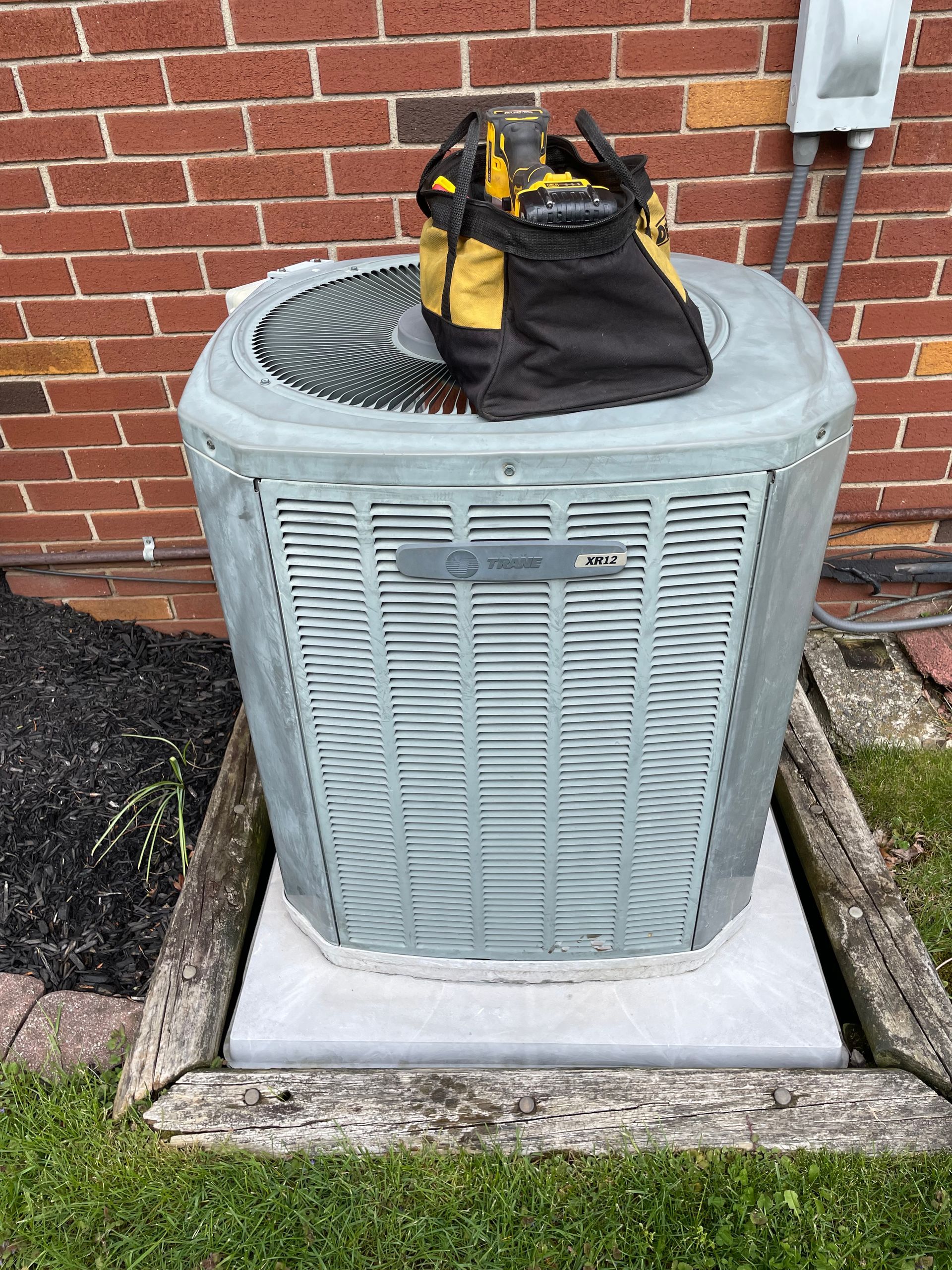 HVAC unit on a concrete pad. Brick wall backdrop with a tool bag resting on top.