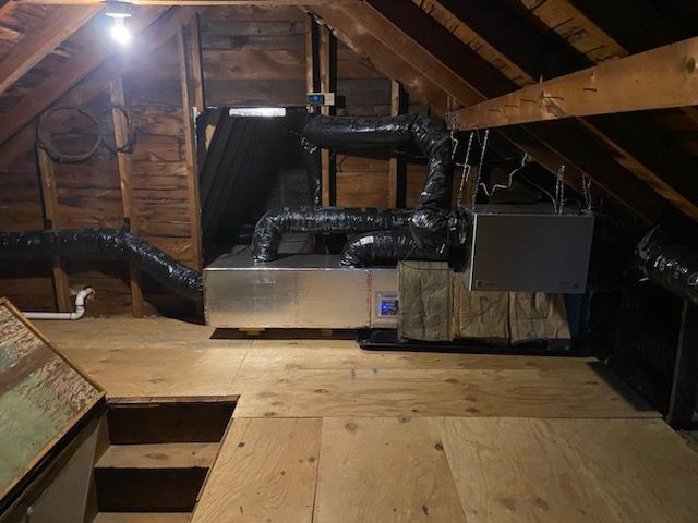 HVAC unit in an attic, with ductwork, plywood flooring, and wooden rafters.