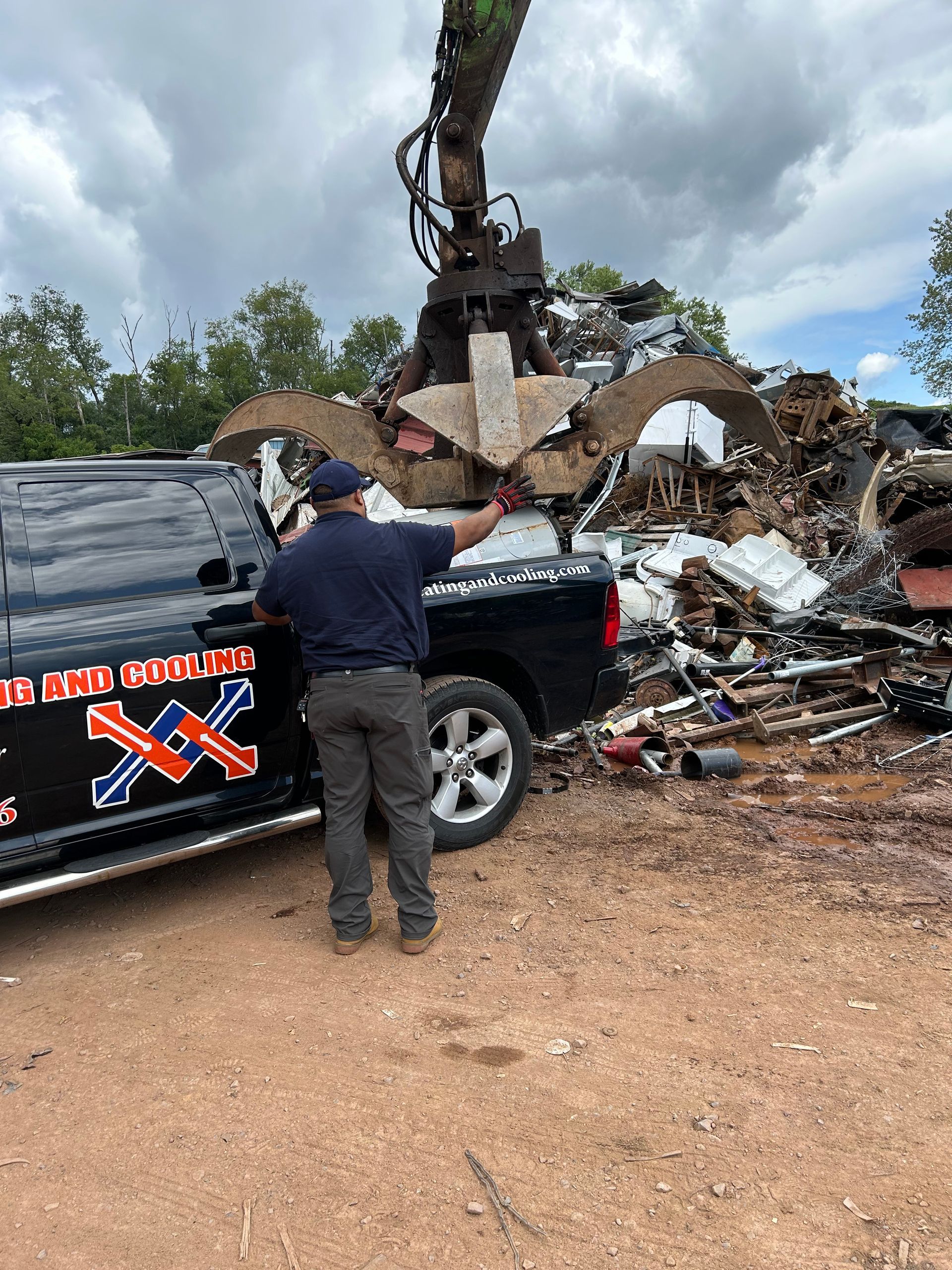 Man gestures toward a pile of scrap metal being moved by a claw, near a black truck, outdoors.