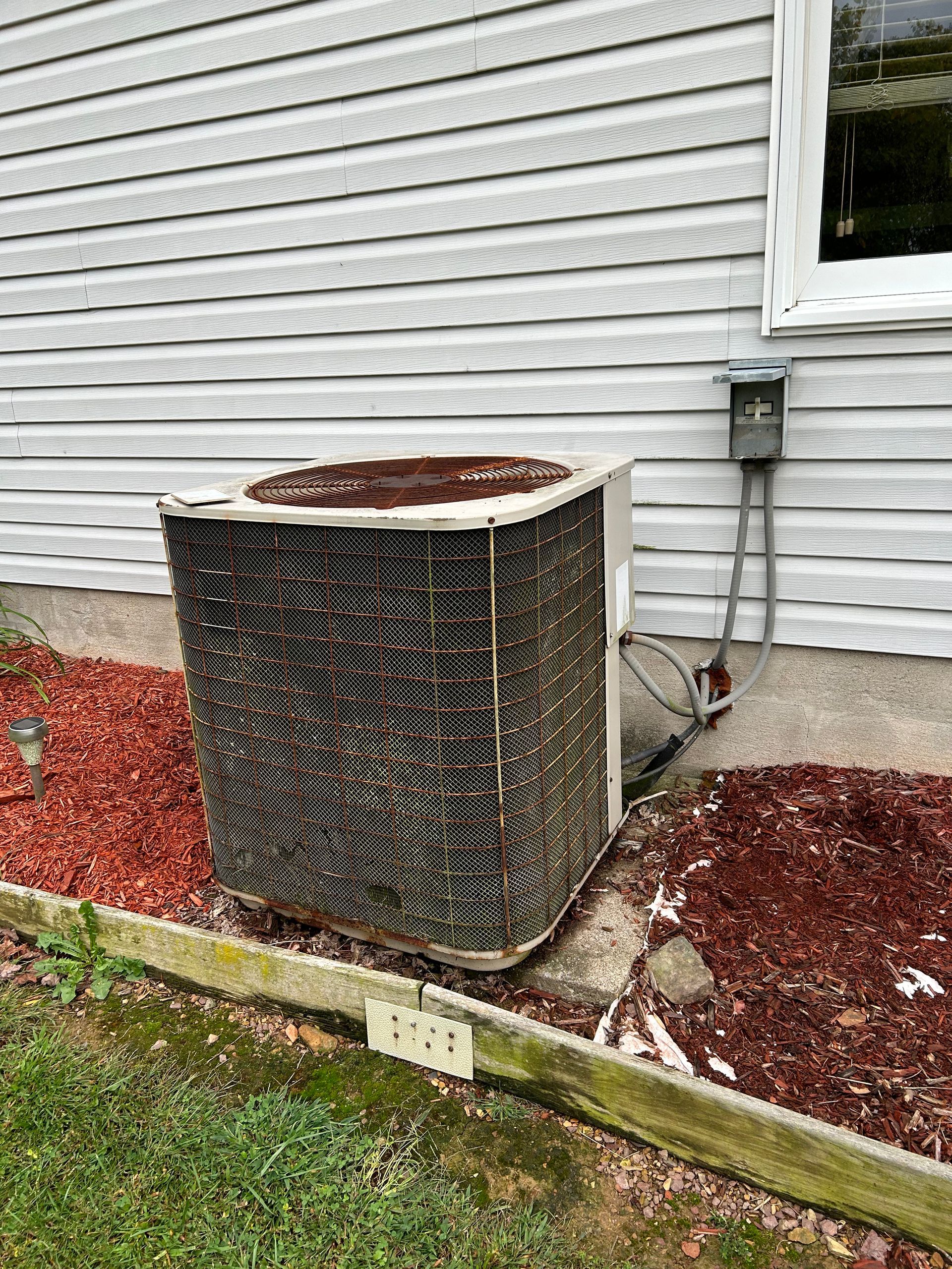 Air conditioner unit outside a house, surrounded by red mulch.