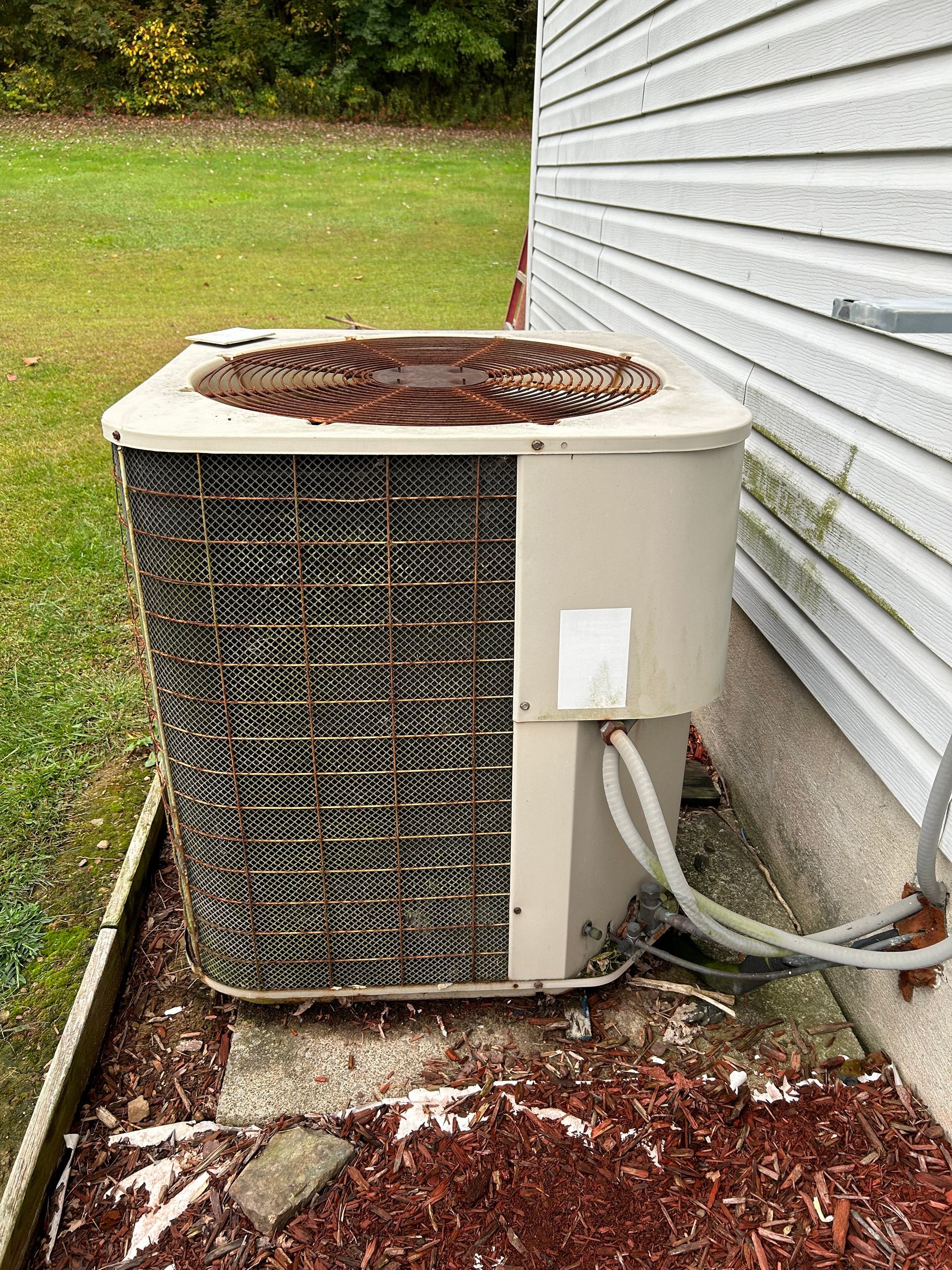 An old, rusty air conditioning unit next to a house with siding, on mulch and grass.