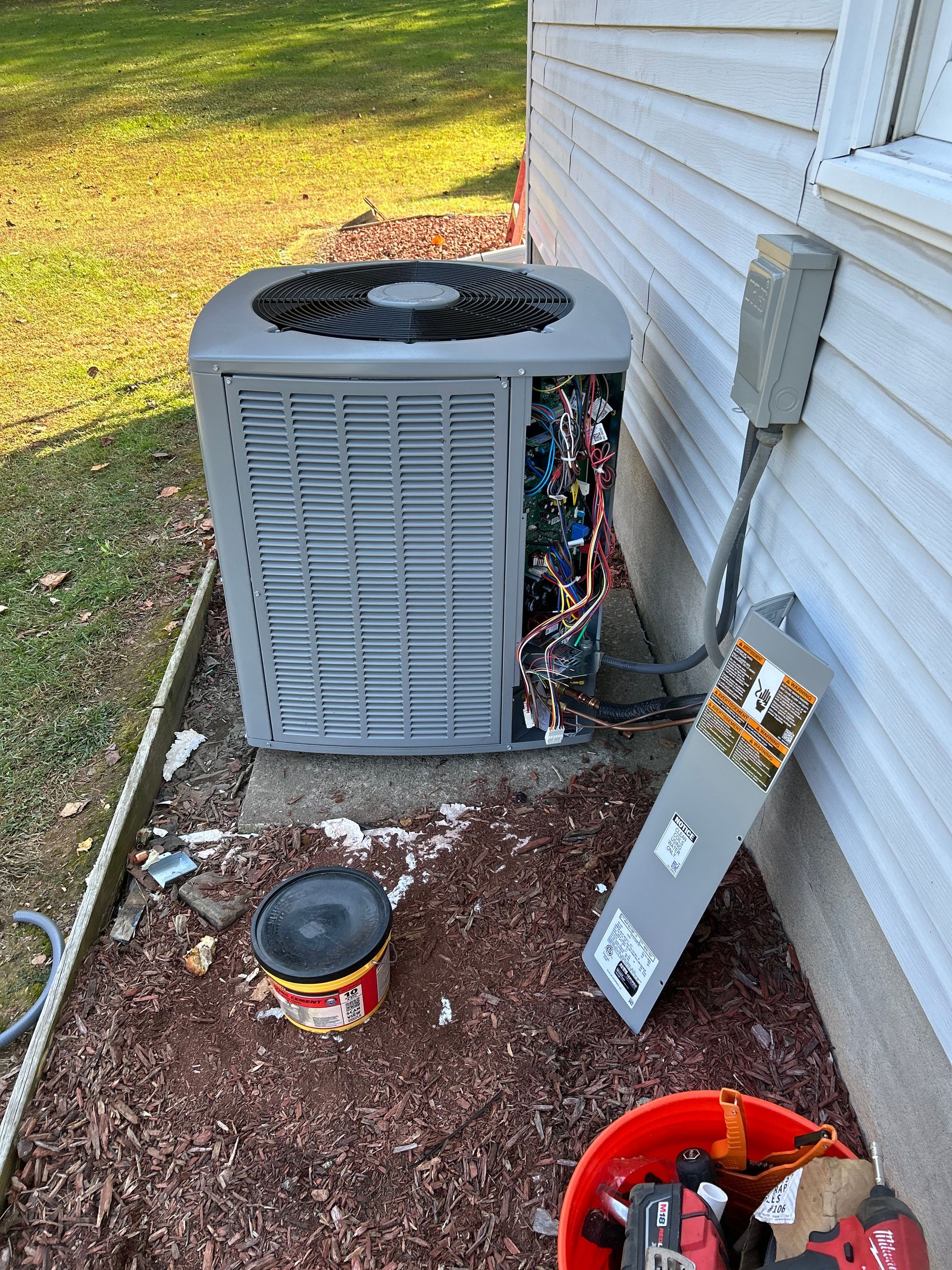 HVAC unit with open side, next to a house. A bucket of tools and paint can sit nearby.