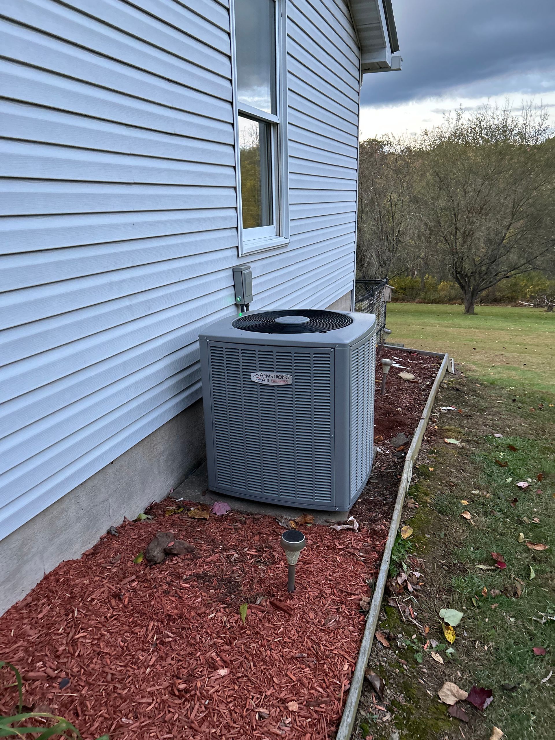 Air conditioning unit next to a house with light blue siding, set in mulch.
