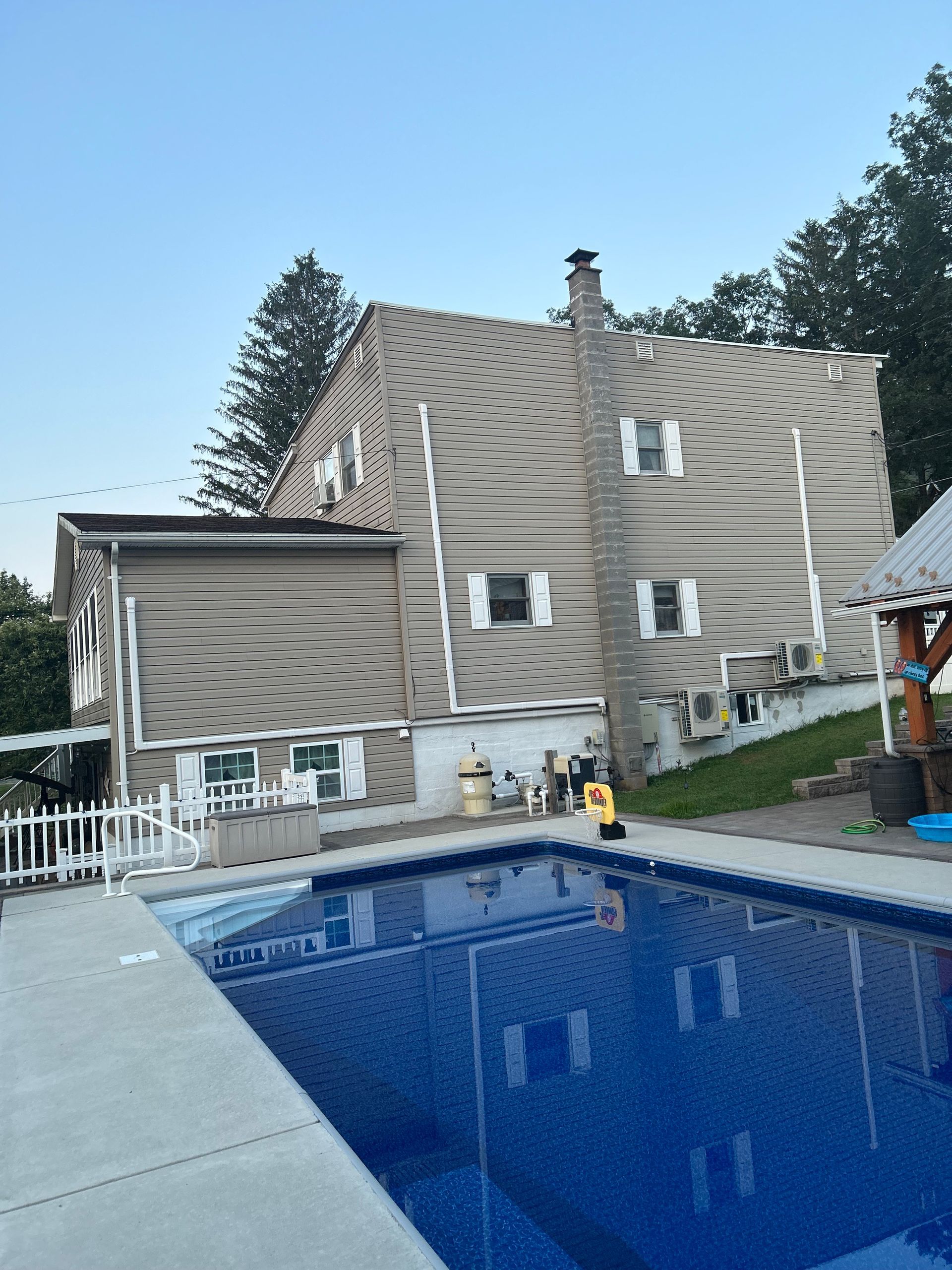 Back of a two-story house with a pool in the foreground. Tan siding, a chimney, and white trim. Blue water.