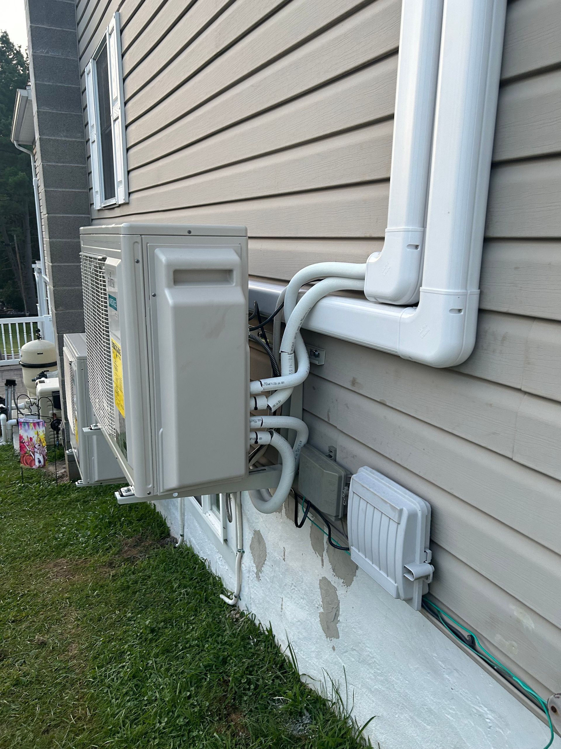 A light gray HVAC unit mounted on a beige house exterior. Pipes and wiring are visible.