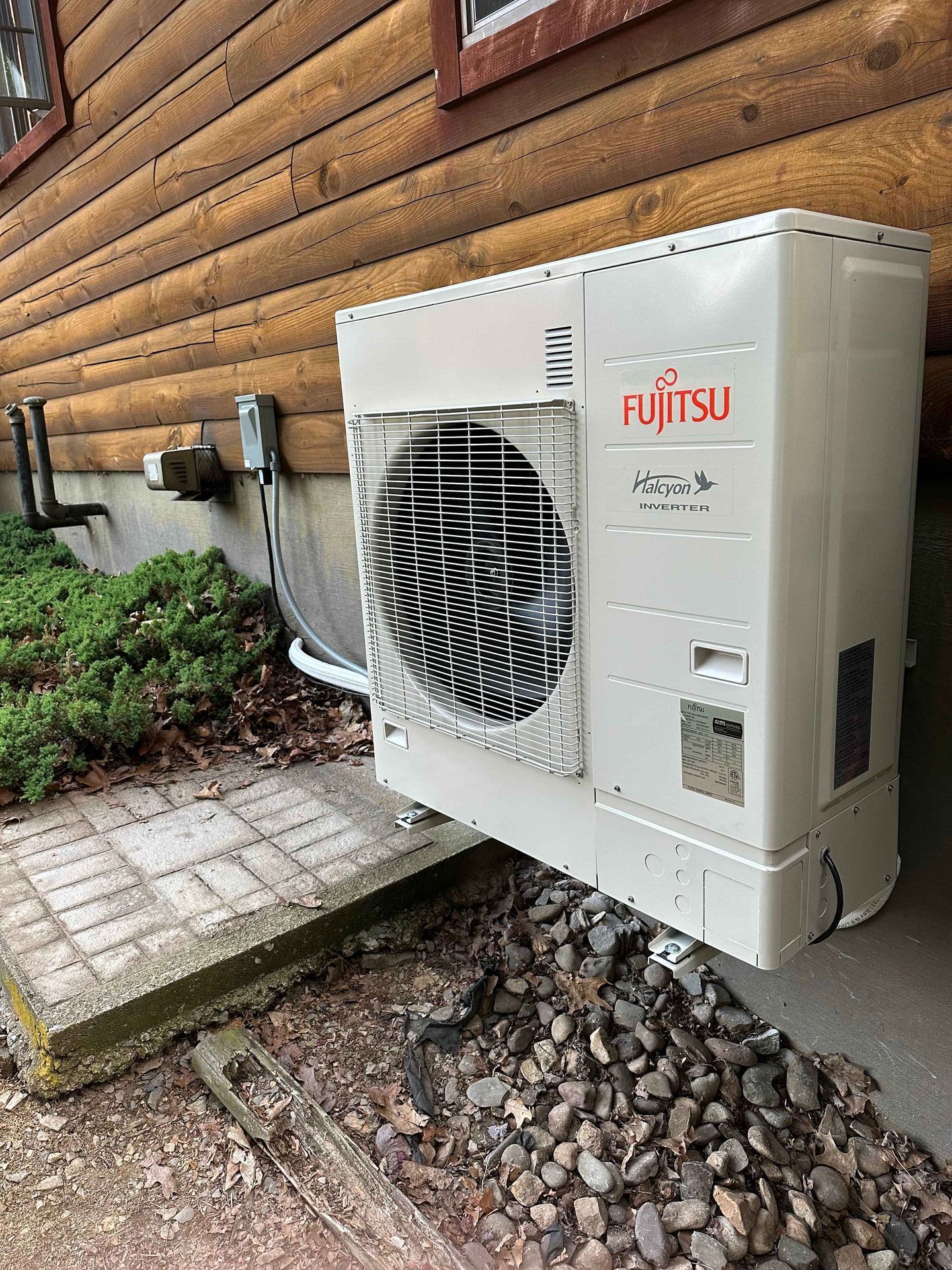 Fujitsu heat pump unit mounted on a wooden building exterior near a small garden.