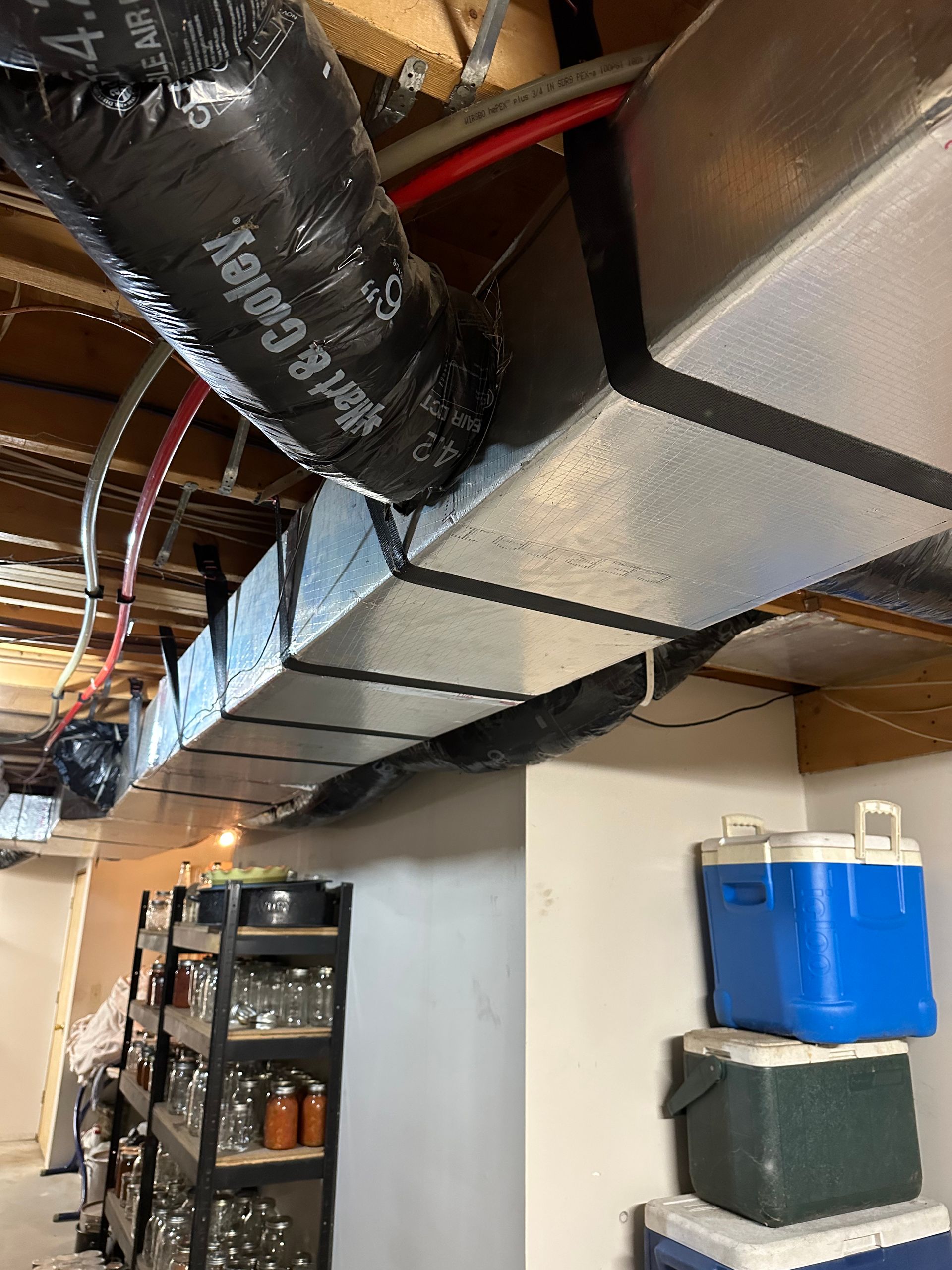 HVAC ductwork in a basement ceiling, with jars on shelves and coolers stacked below.