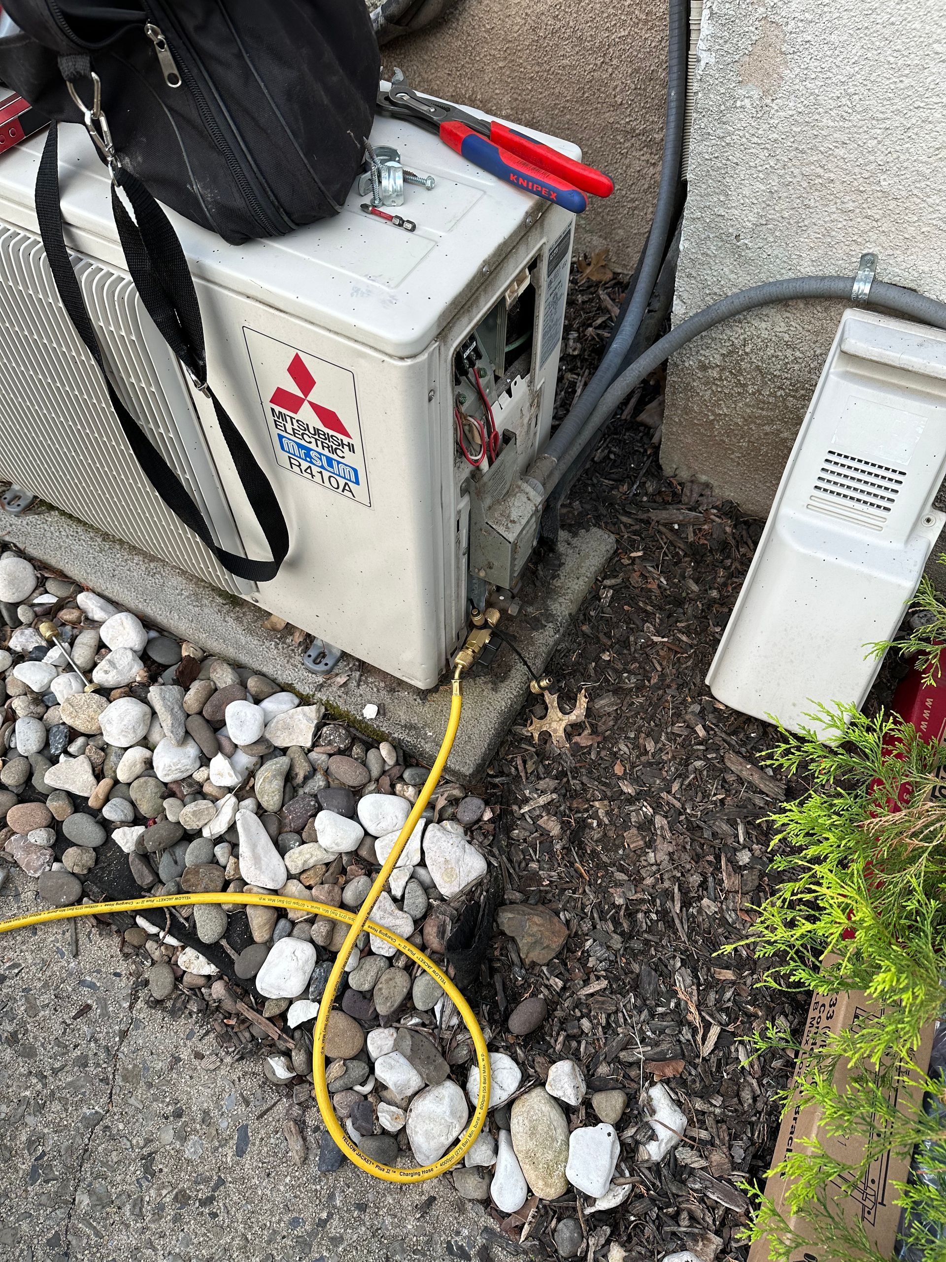 Mitsubishi outdoor AC unit, tools, and wiring, near stone, ground, and a wall.