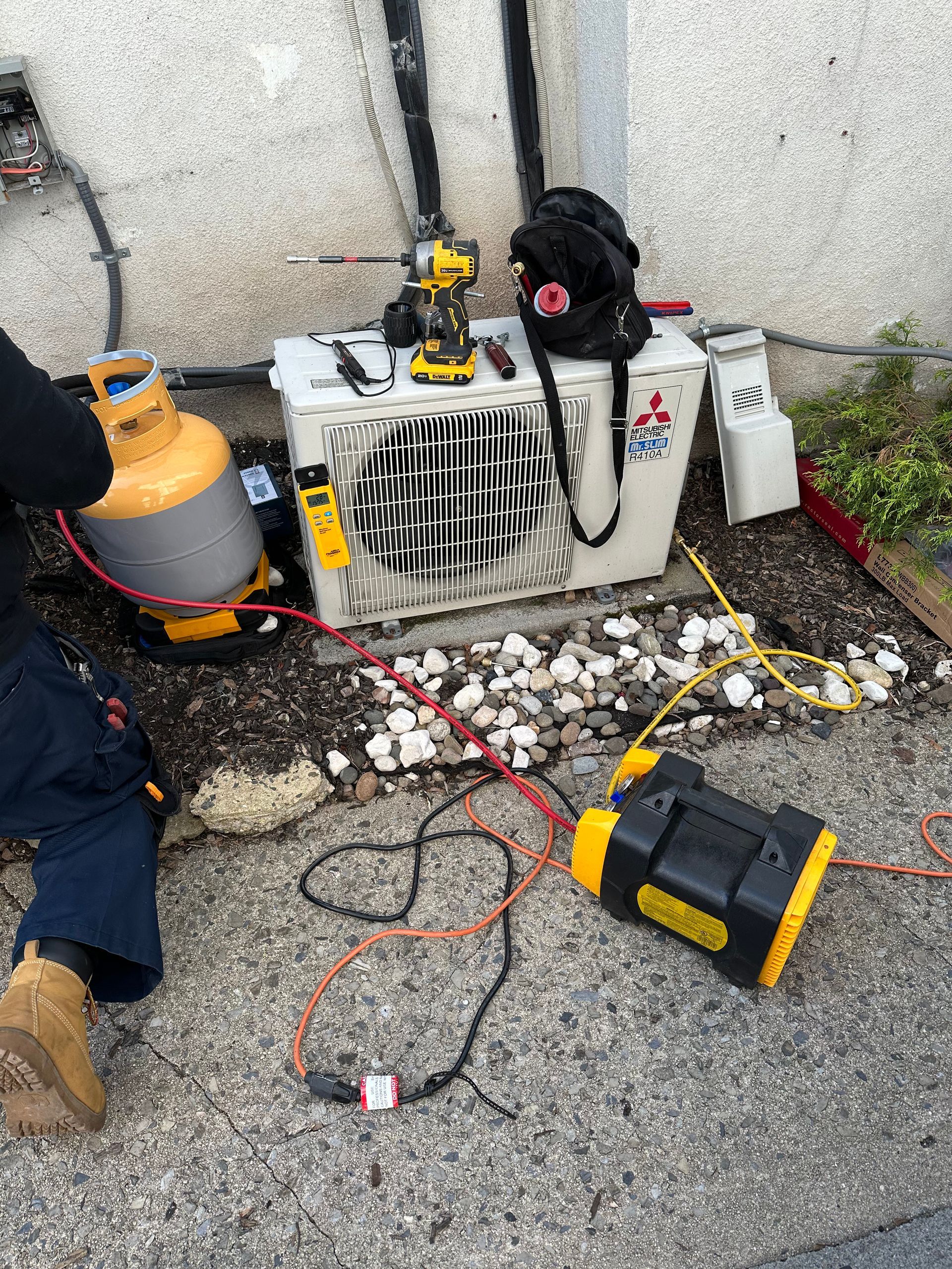 HVAC technician working on an outdoor AC unit with tools, gas tank, and power cords. Concrete ground, gray wall.