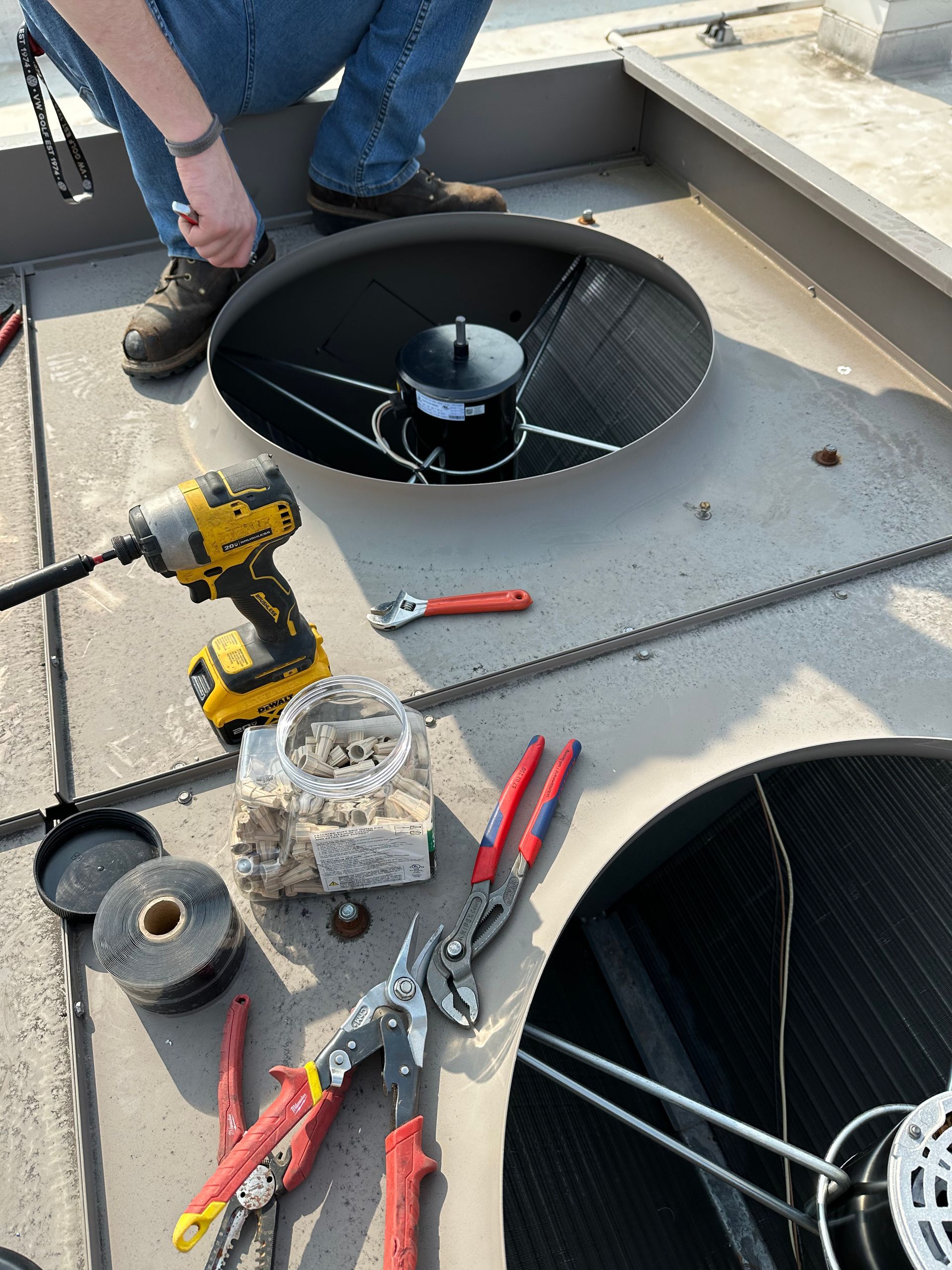 Person on rooftop repairs a black fan. Tools, including a drill and pliers, surround the fan.