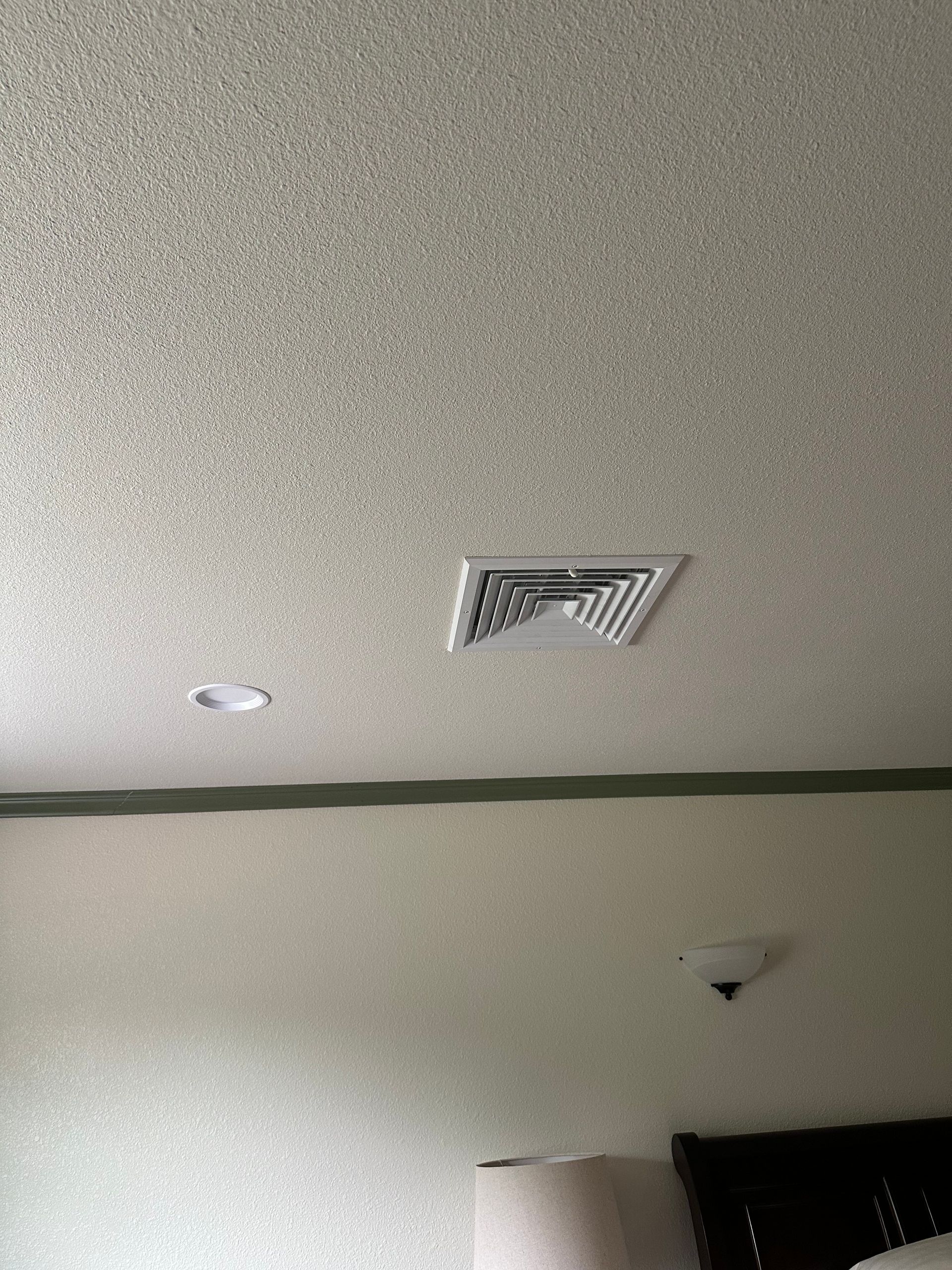 Ceiling with textured finish, a vent, and a recessed light. Below, a wall and a bed are visible.