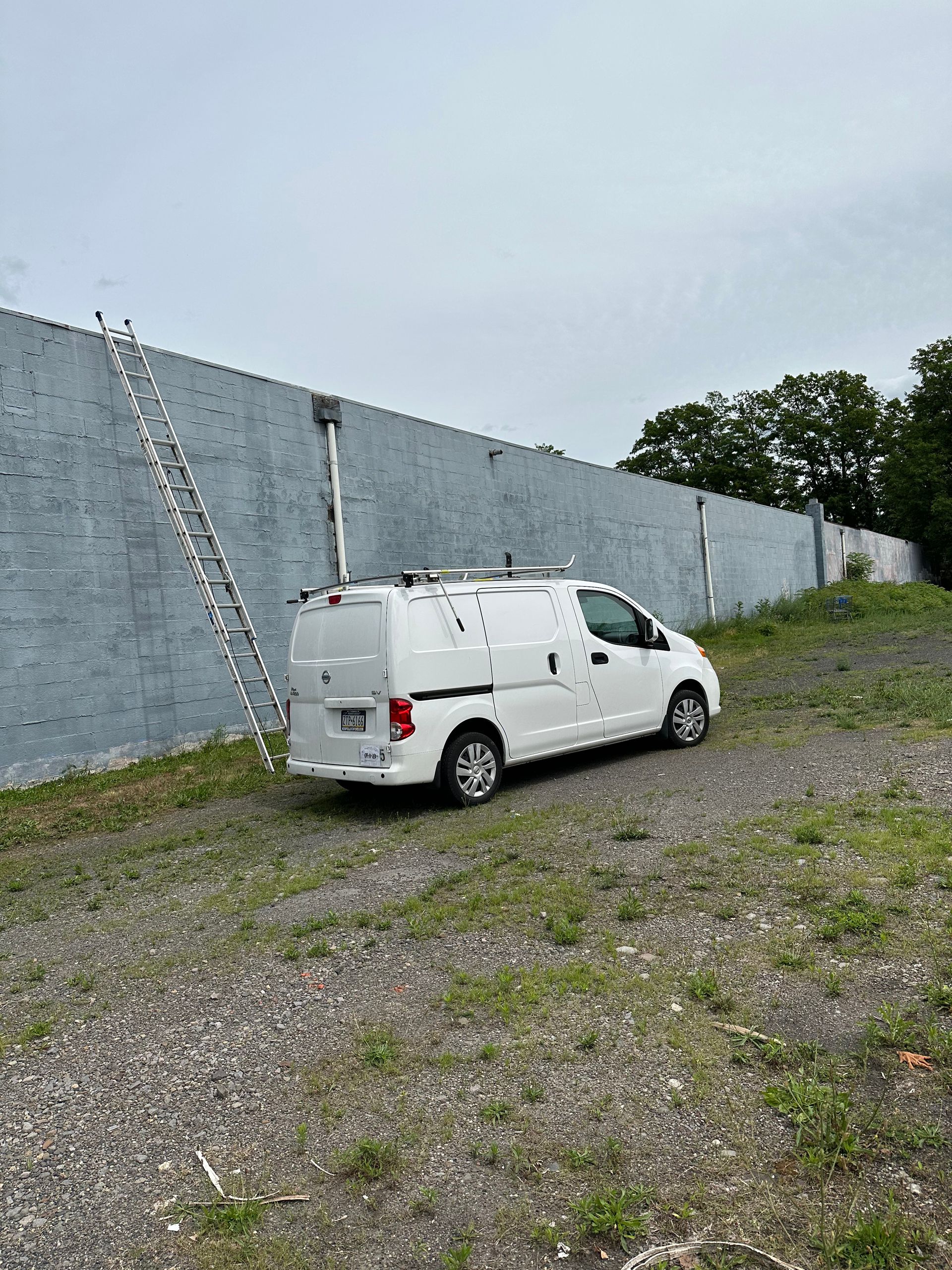 White van parked on gravel next to a tall, gray brick wall with a ladder leaning against it.