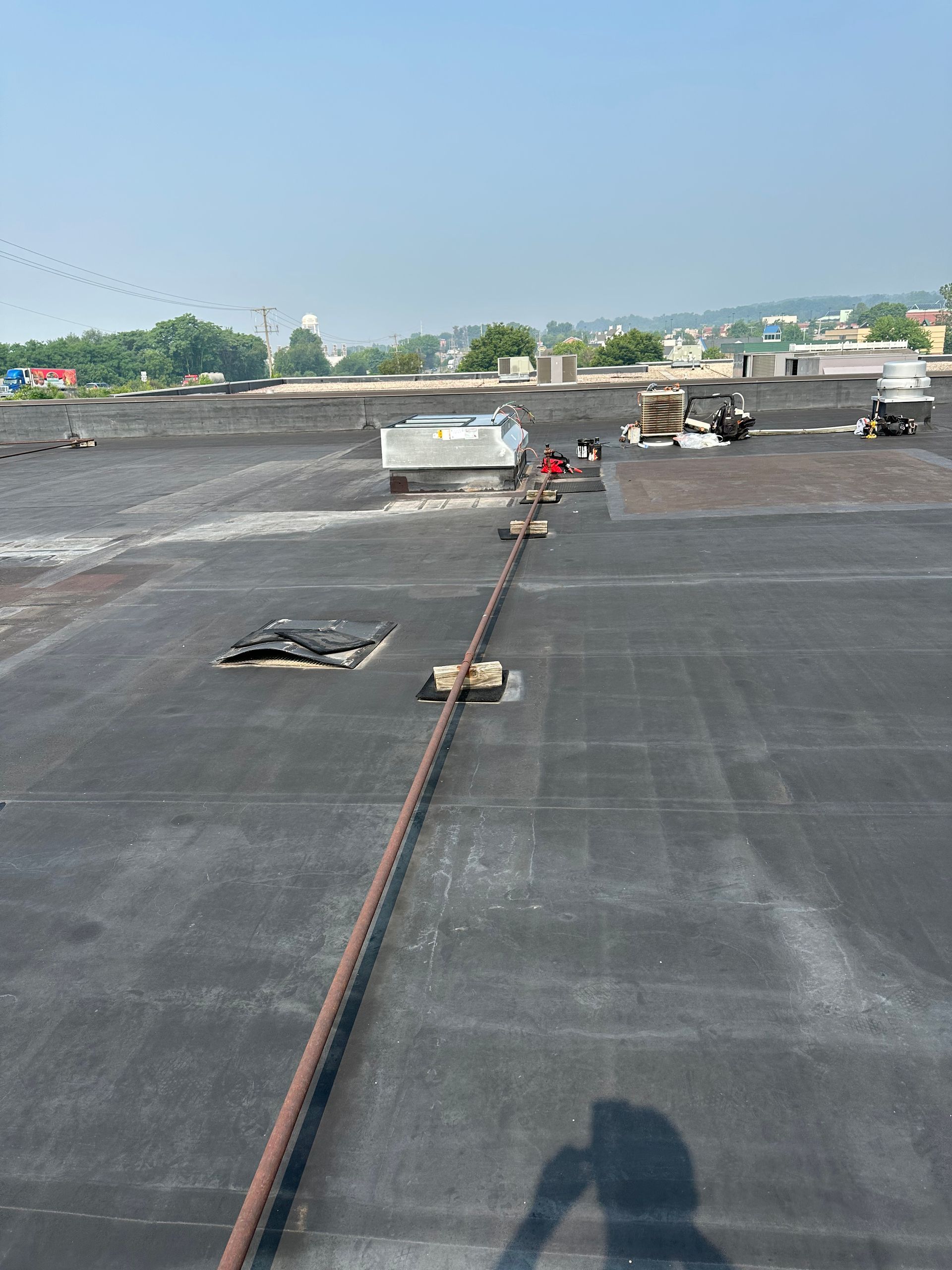 Black rooftop with a safety cable, equipment, and distant buildings under a blue sky.