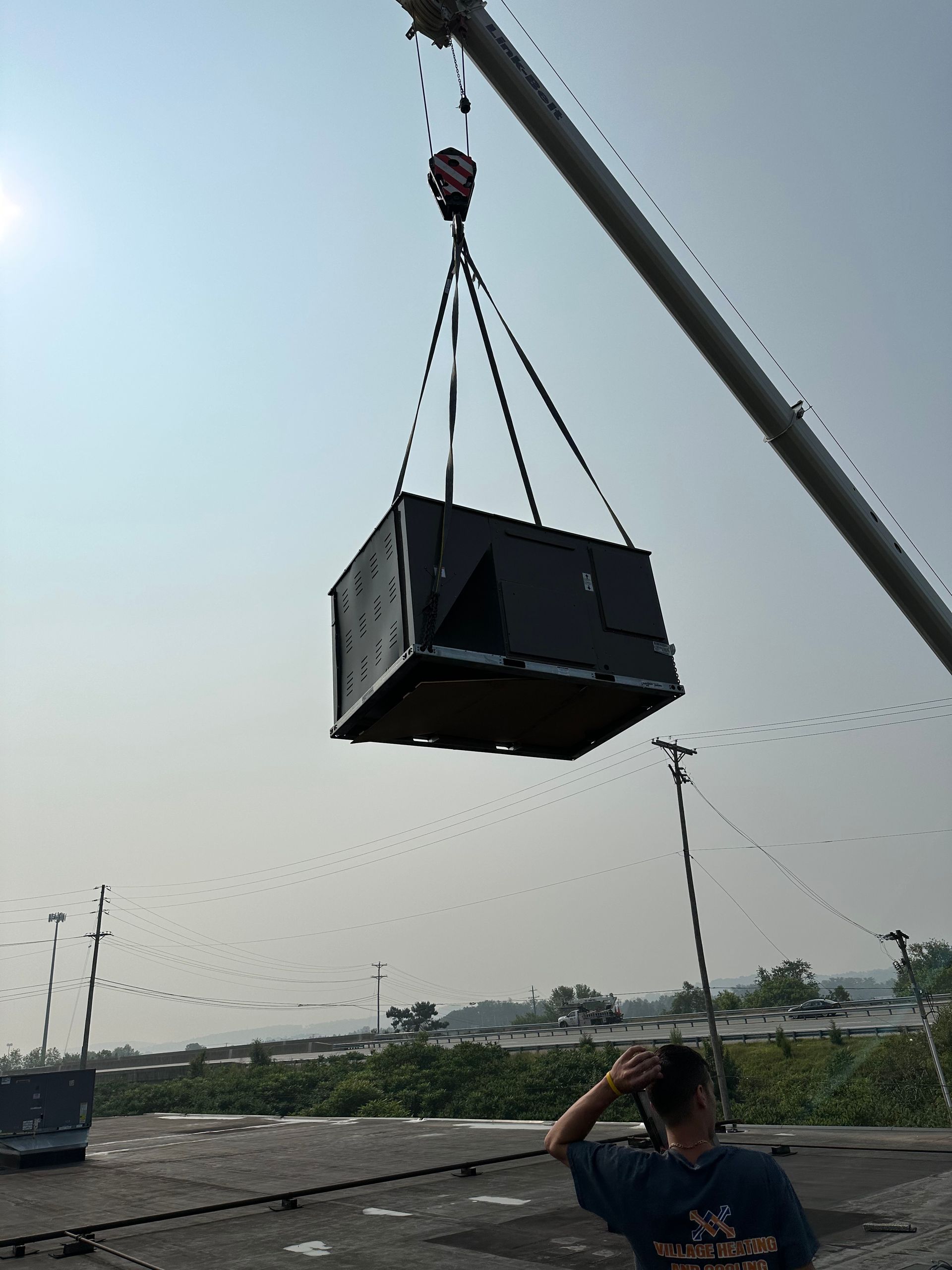 A crane lifting a large, dark box. A person watches. Outdoors, bright sky.