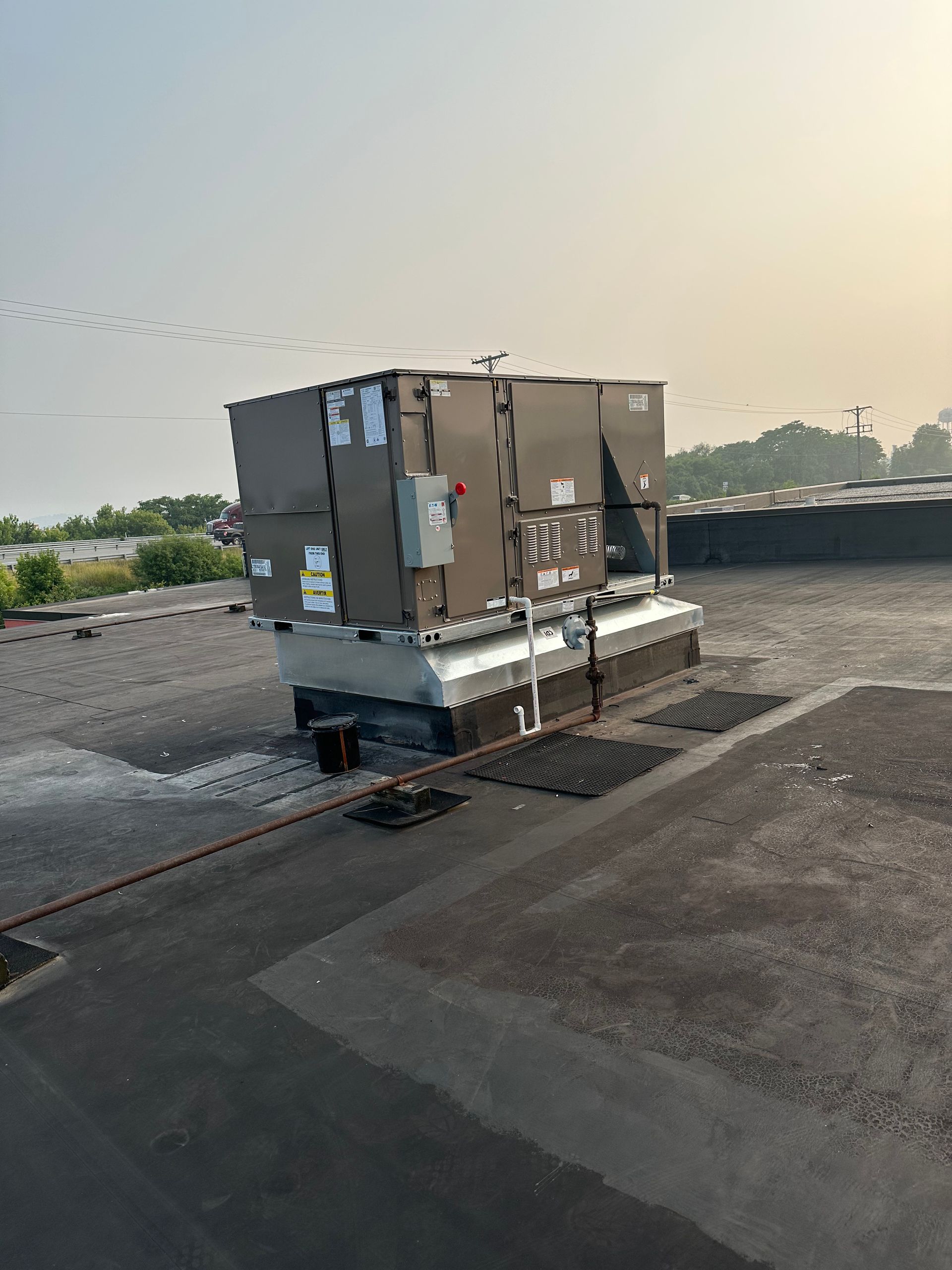 Rooftop HVAC unit, metallic gray, on a black flat roof, cloudy sky background.