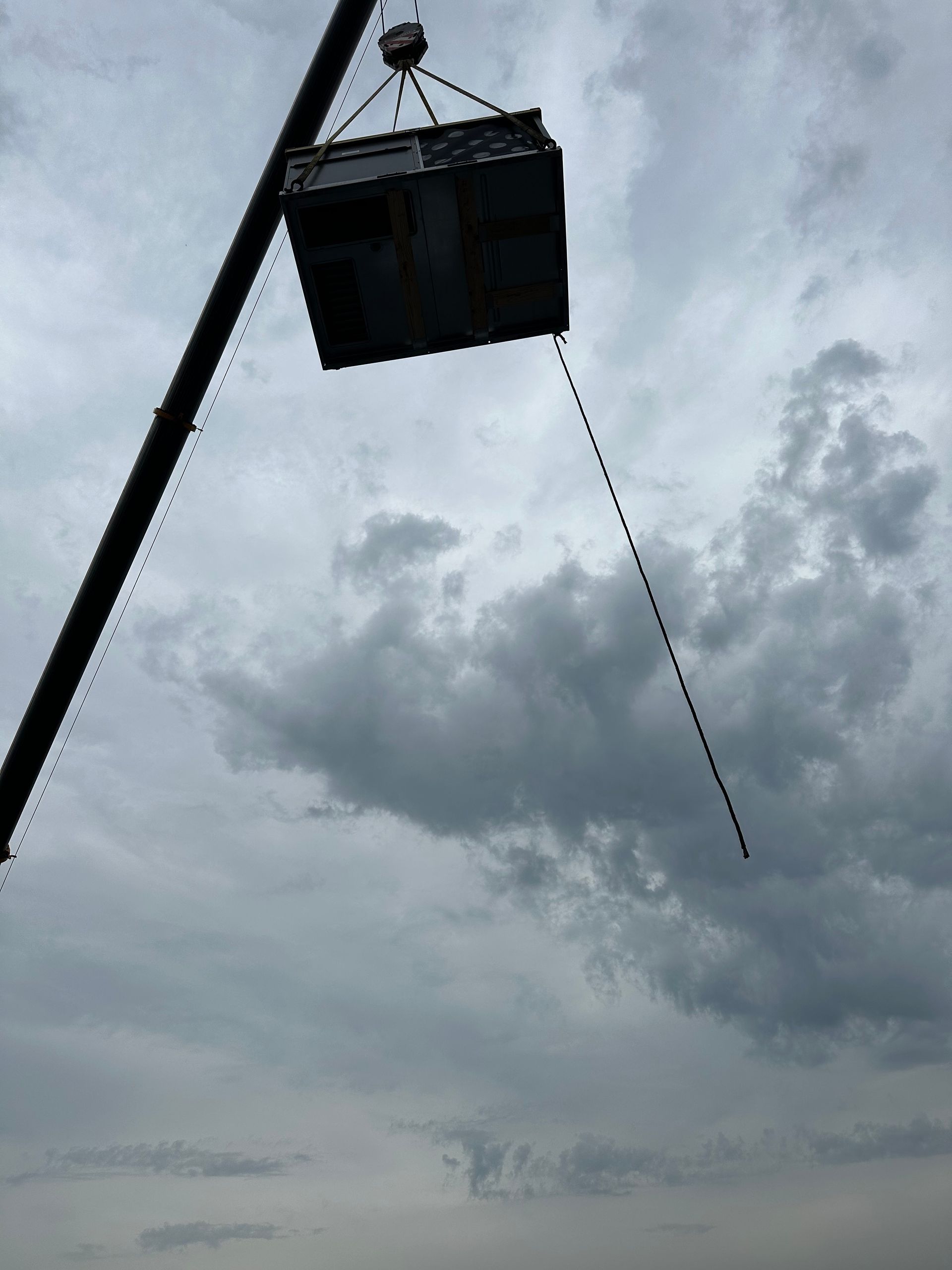A large, square metal object suspended by a crane against a cloudy sky.