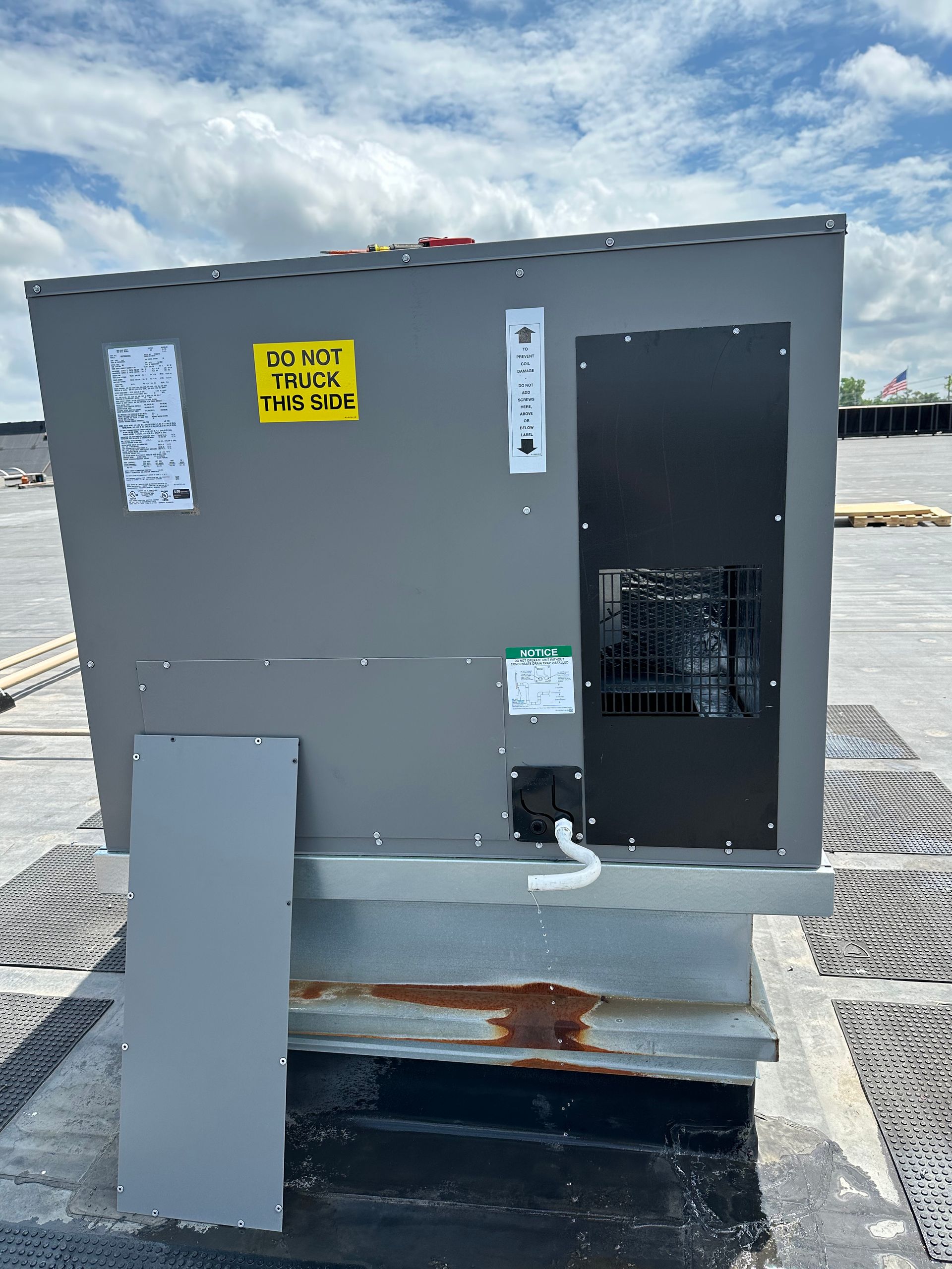 HVAC rooftop unit, gray metal with a black door, mounted on a platform on a flat roof, cloudy sky.