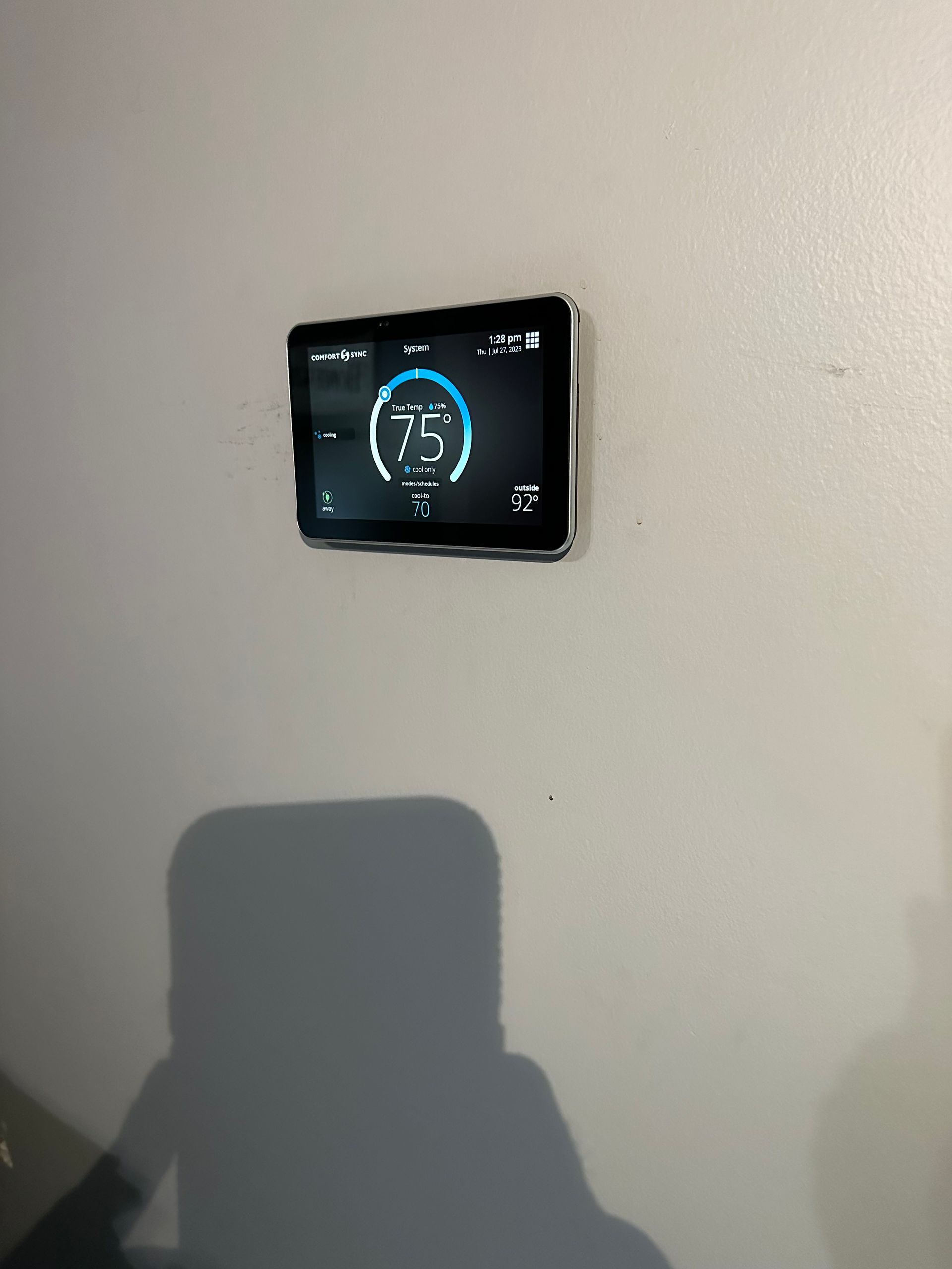 A digital thermostat on a wall displaying 75 degrees. The shadow of a person is in the foreground.