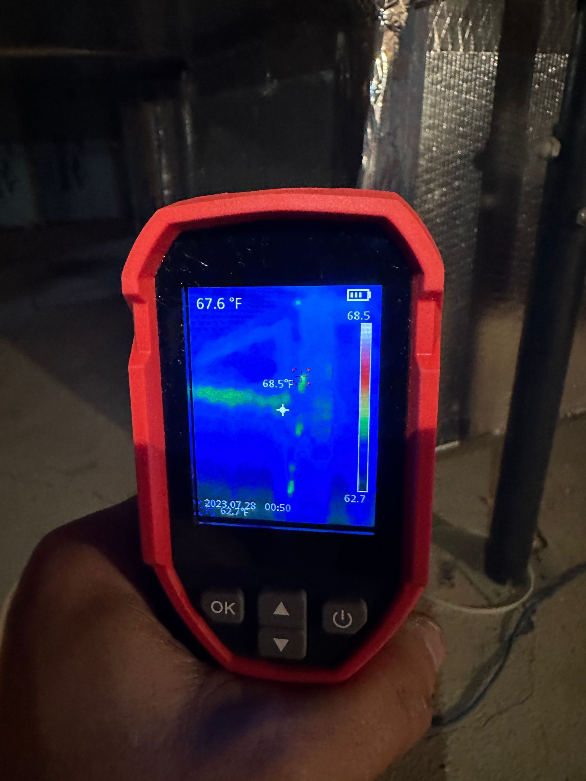 Infrared camera displaying a thermal image of a dark space, held by a person; temperature readings are visible.