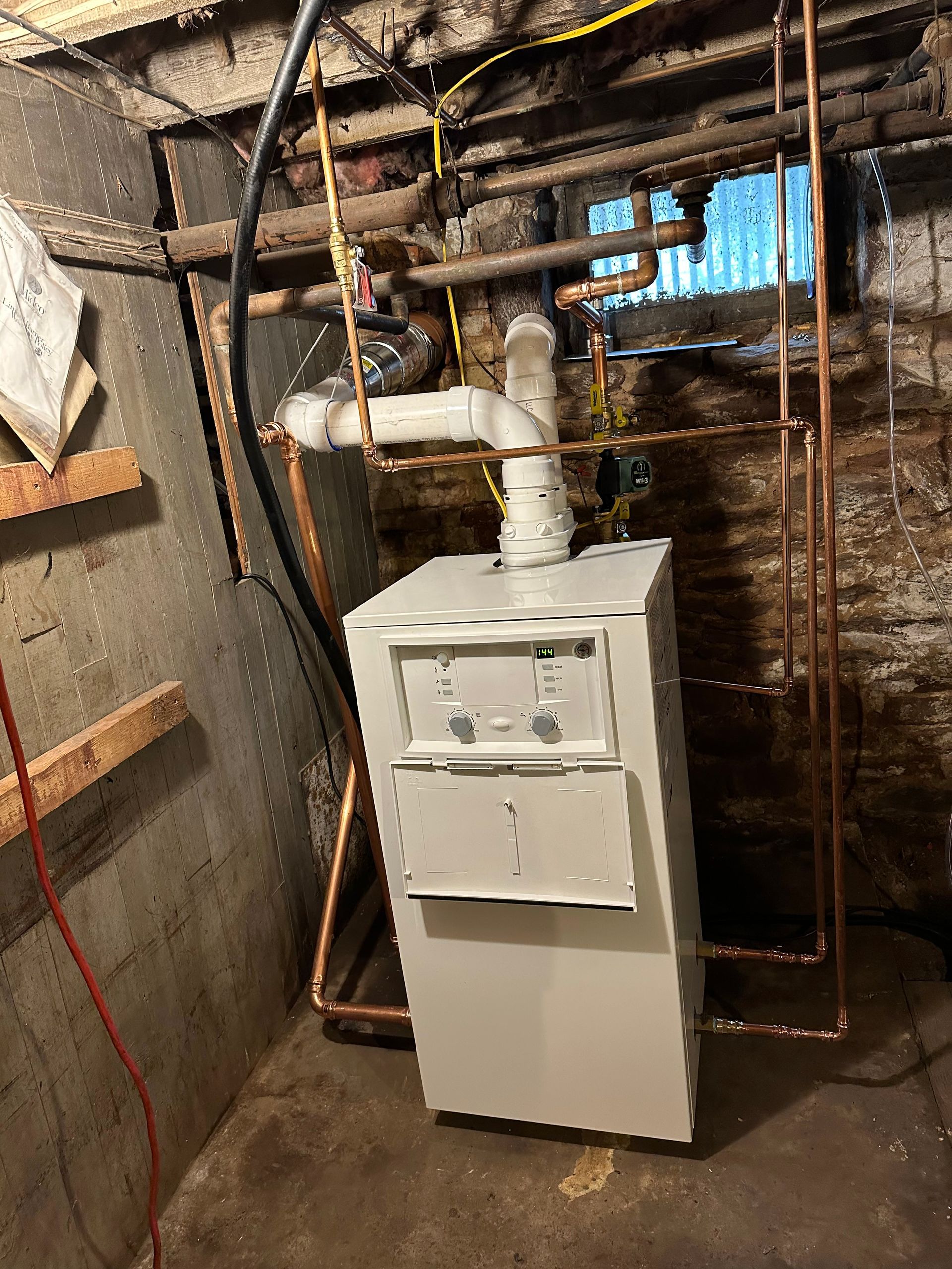 A white furnace in a basement, with copper pipes. The walls are brick.