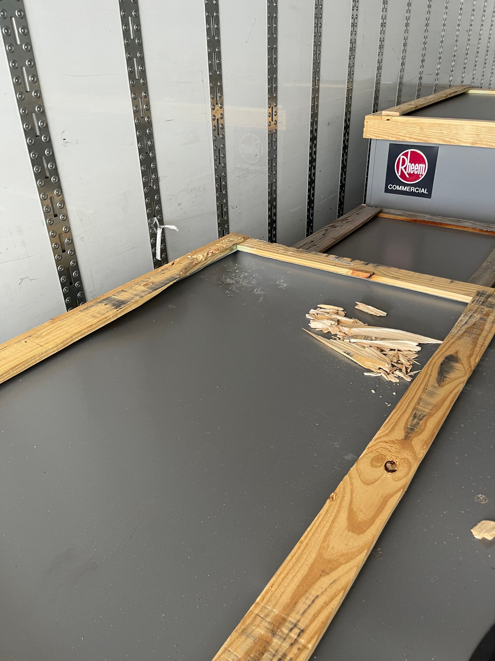 Inside a cargo trailer, a wooden frame with debris on a gray surface next to a container.