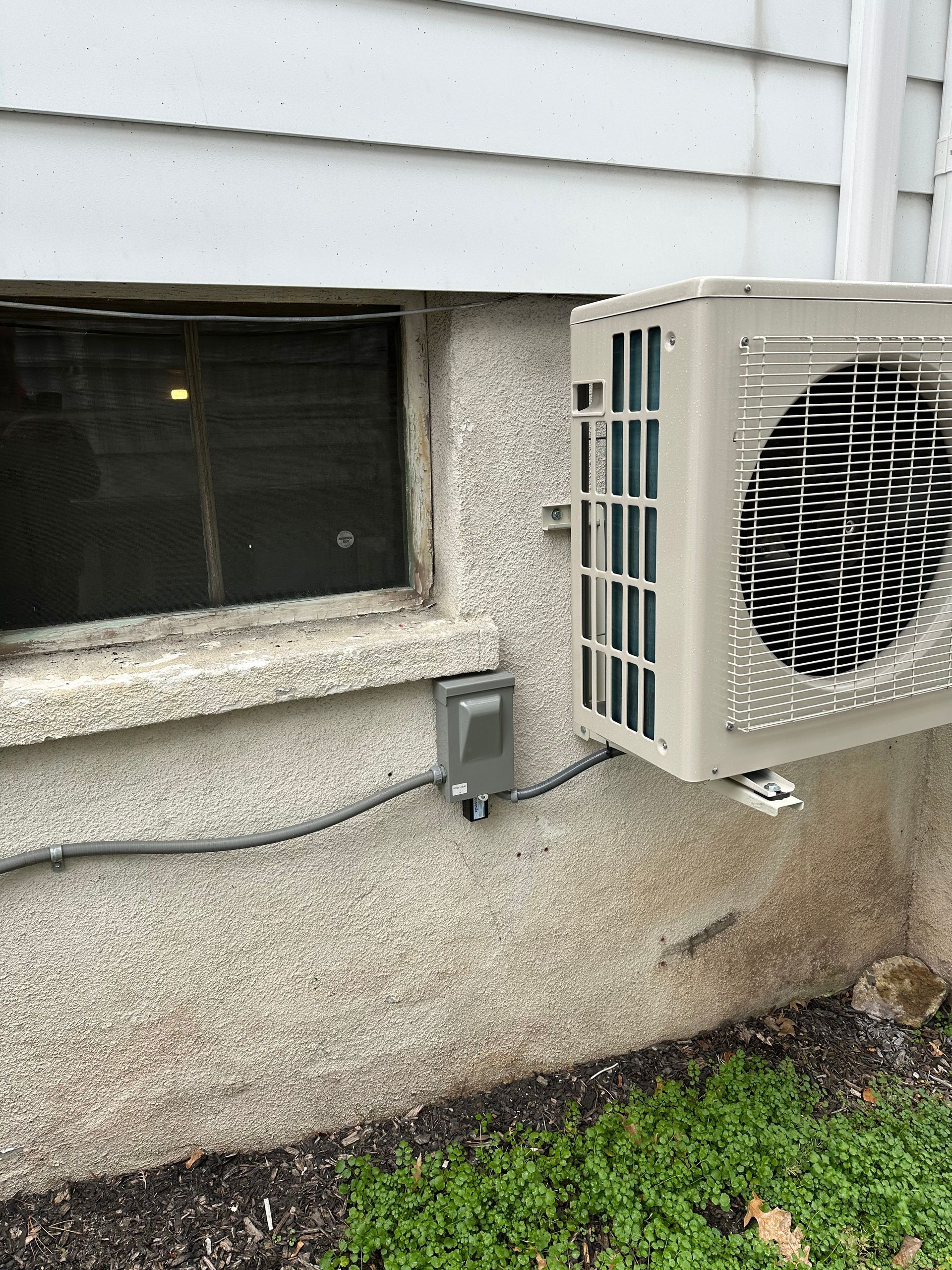A tan air conditioning unit mounted on a stucco wall next to a small window.