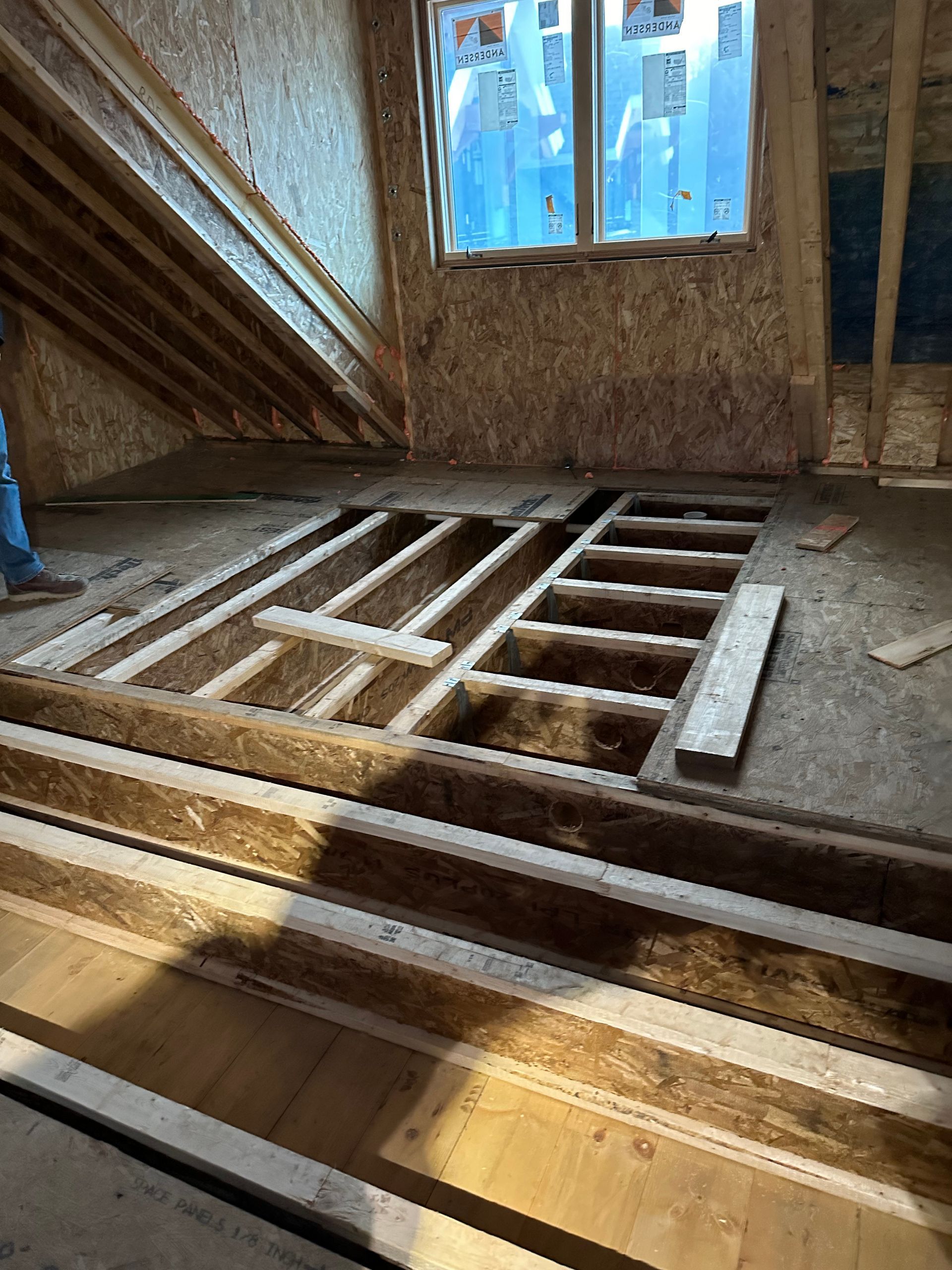 Interior of a room under construction with exposed wooden beams, a window, and unfinished flooring.