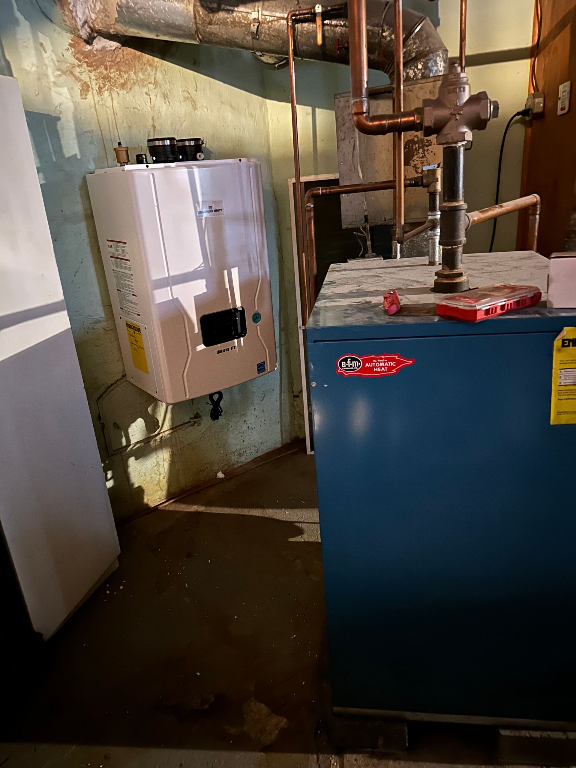 A white tankless water heater mounted on a green basement wall, next to a blue furnace.