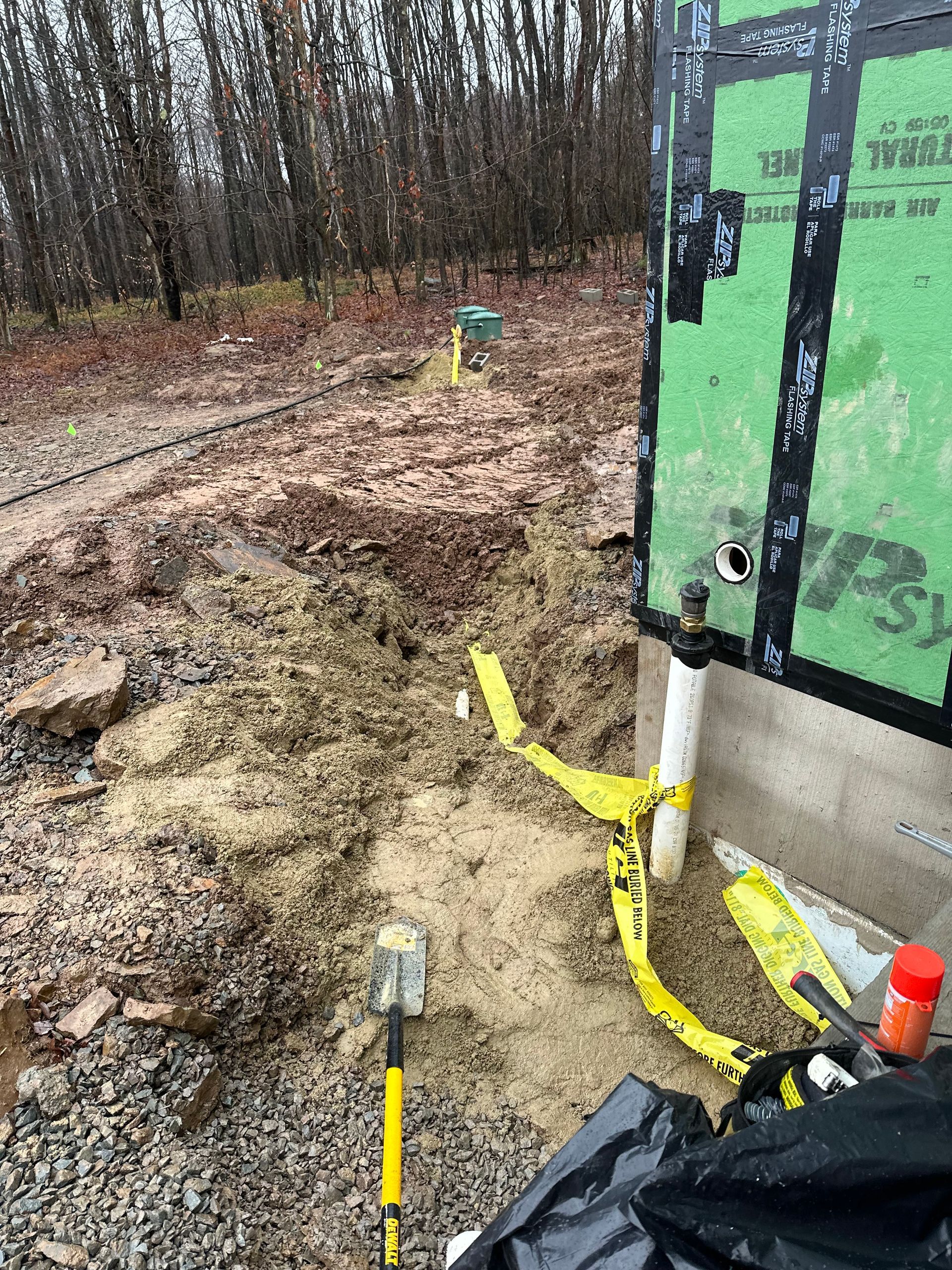 Construction site with muddy ground, exposed pipes marked with yellow tape, near a building foundation.