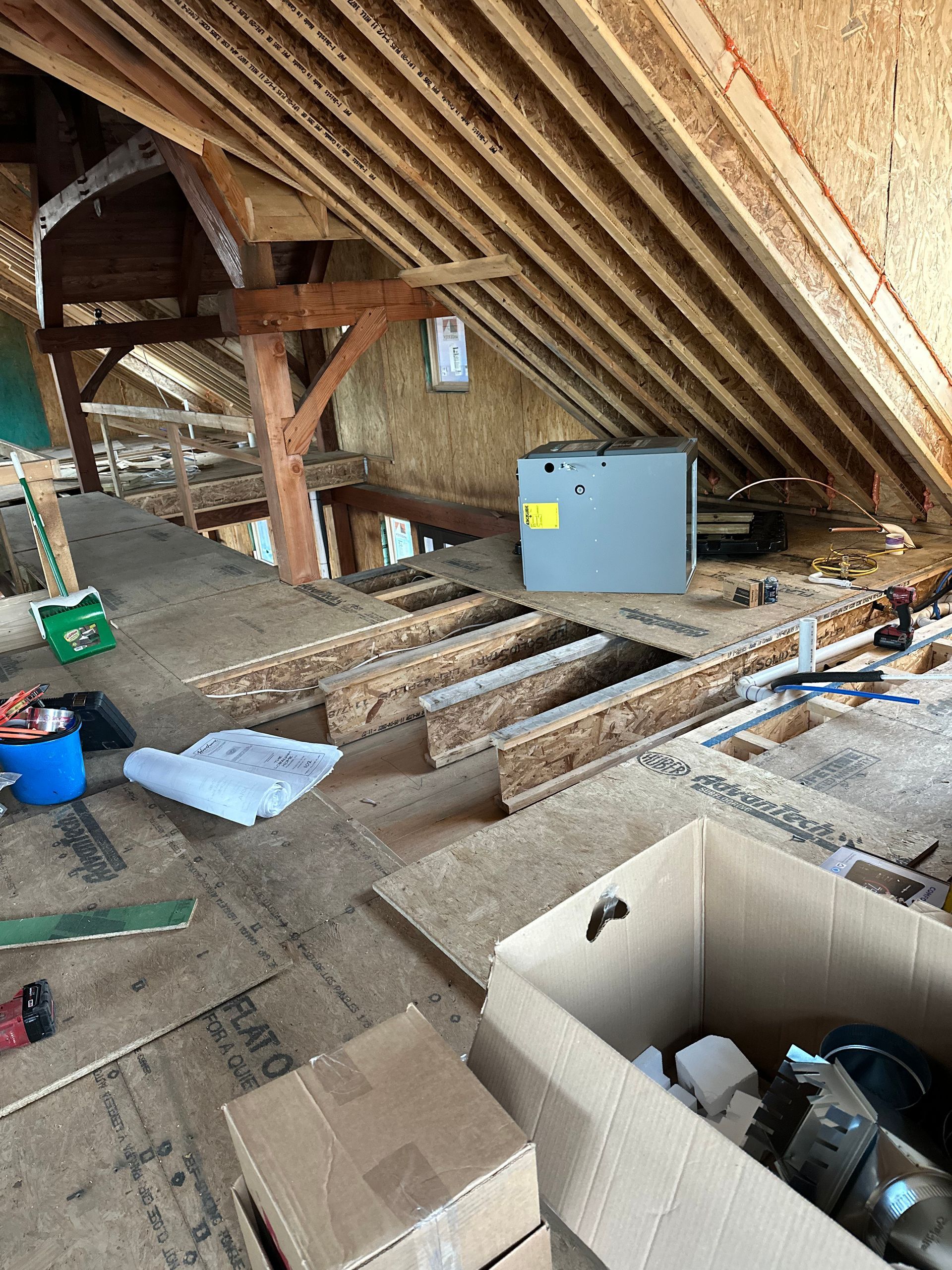Interior view of an unfinished attic. Wood framing, HVAC unit, boxes, and tools are visible.