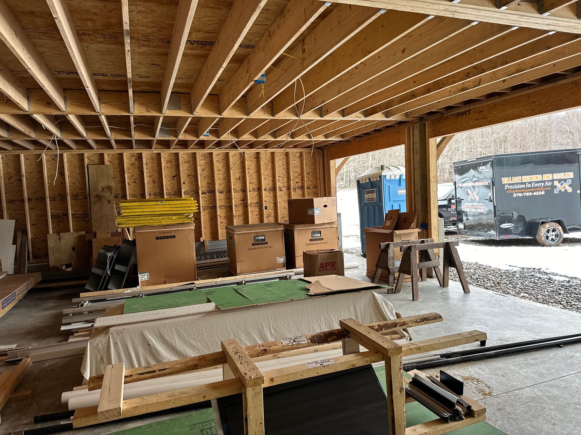 Construction site: unfinished wooden structure with boxes, tools, and a porta-potty outside; overcast day.