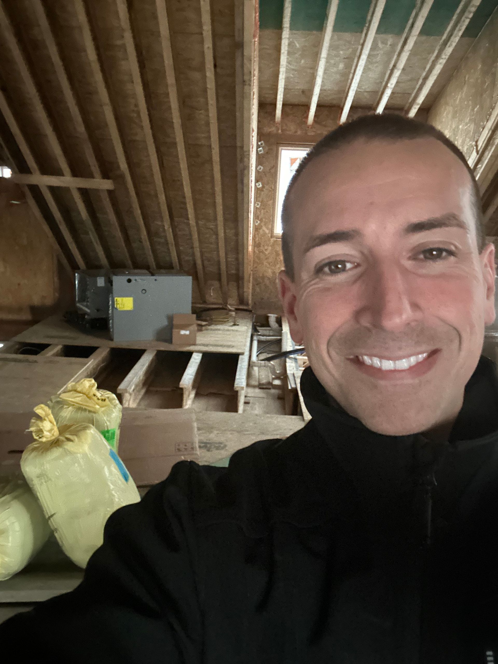 Man smiles in a wood-framed attic; insulation bags, HVAC unit, and rough flooring are visible.