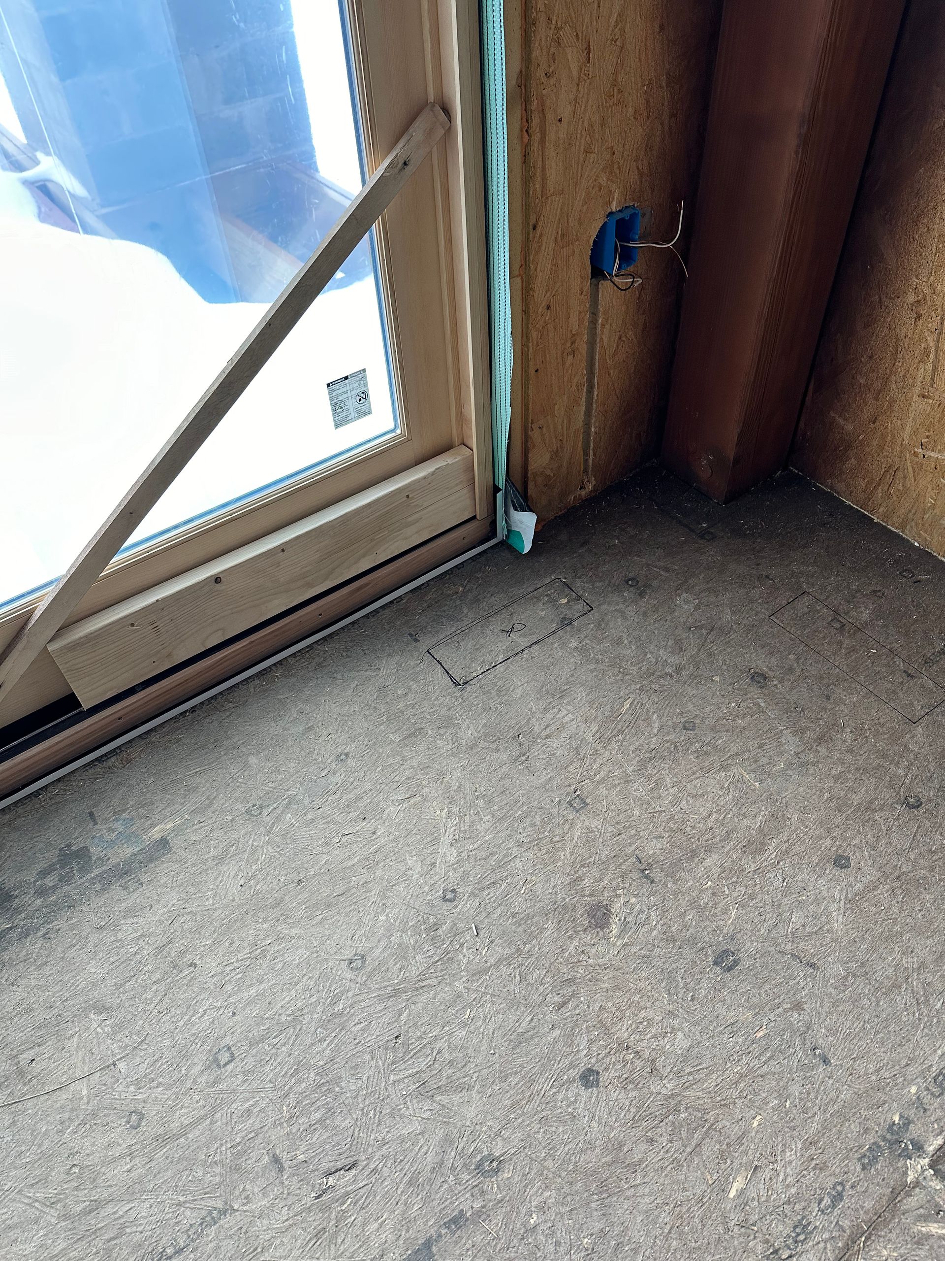 Close-up of window and floor; dark floor with cracks, pale wood window frame, blue electrical box on wall.