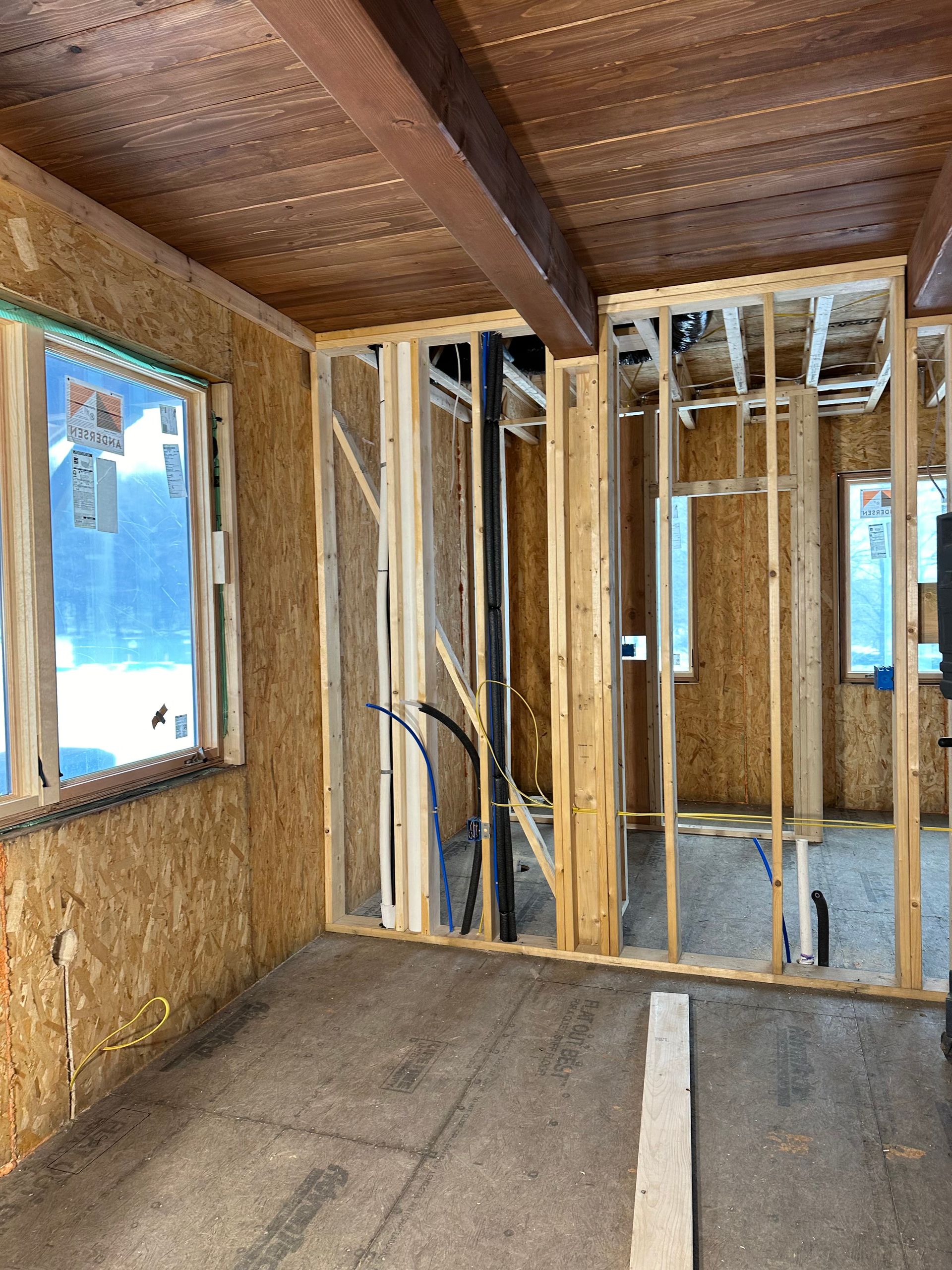 Interior construction view. Bare wooden framing, exposed pipes, and OSB walls in a room with a window and exposed beams.