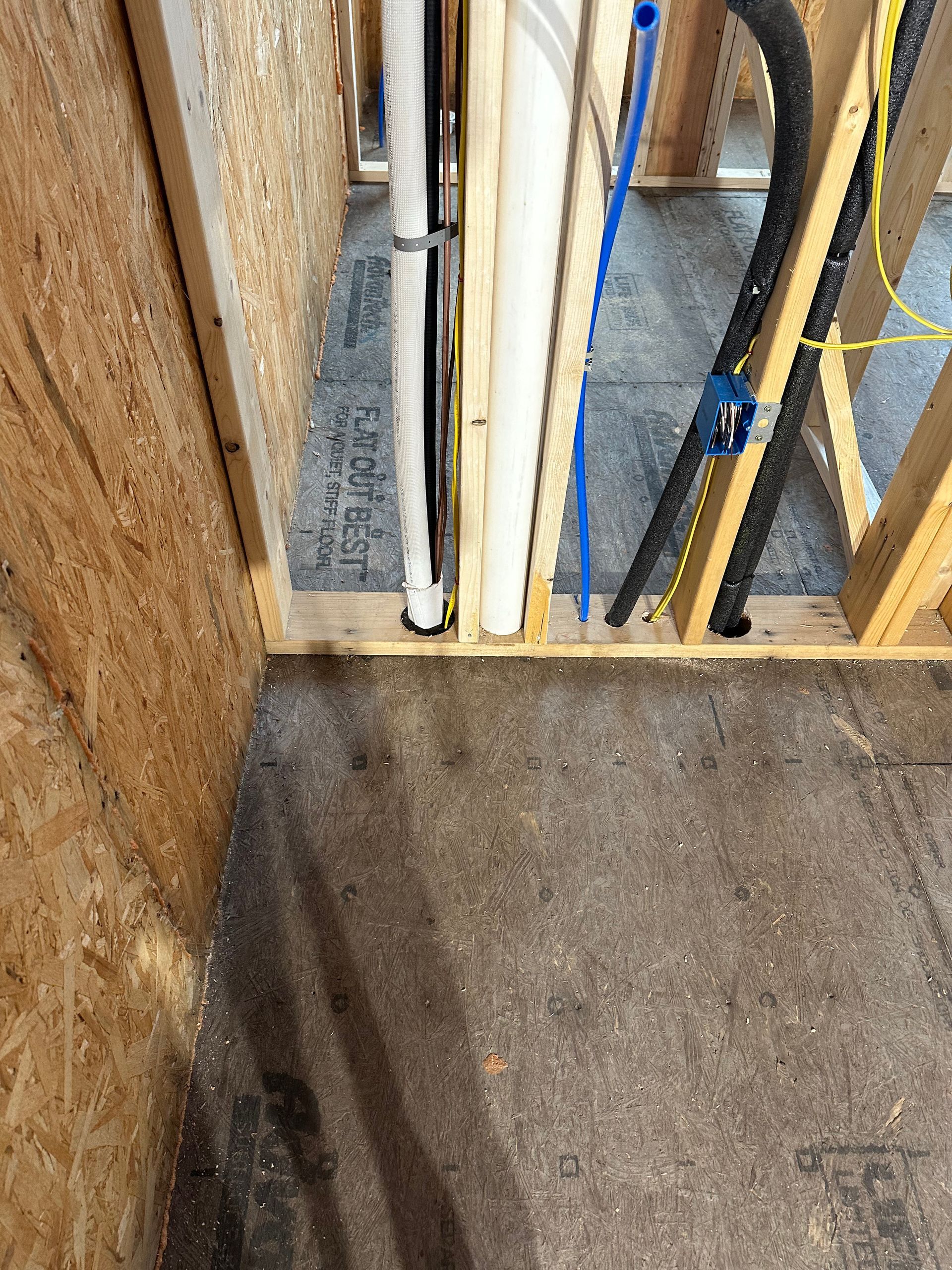 Interior view of exposed pipes and wires within wooden wall framing. Gray flooring.