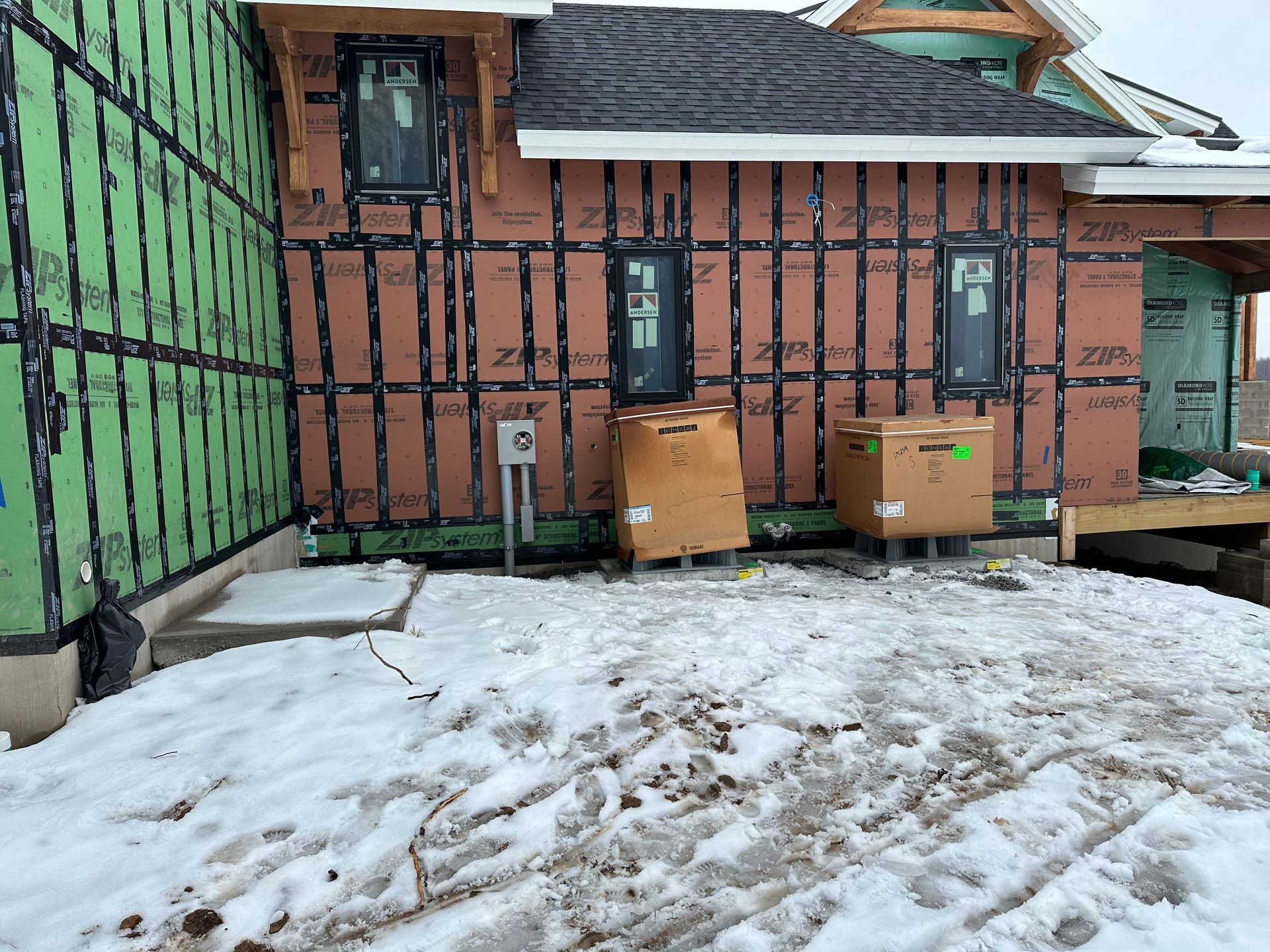 Construction site exterior: house with sheathing, windows, cardboard boxes on snow-covered ground.