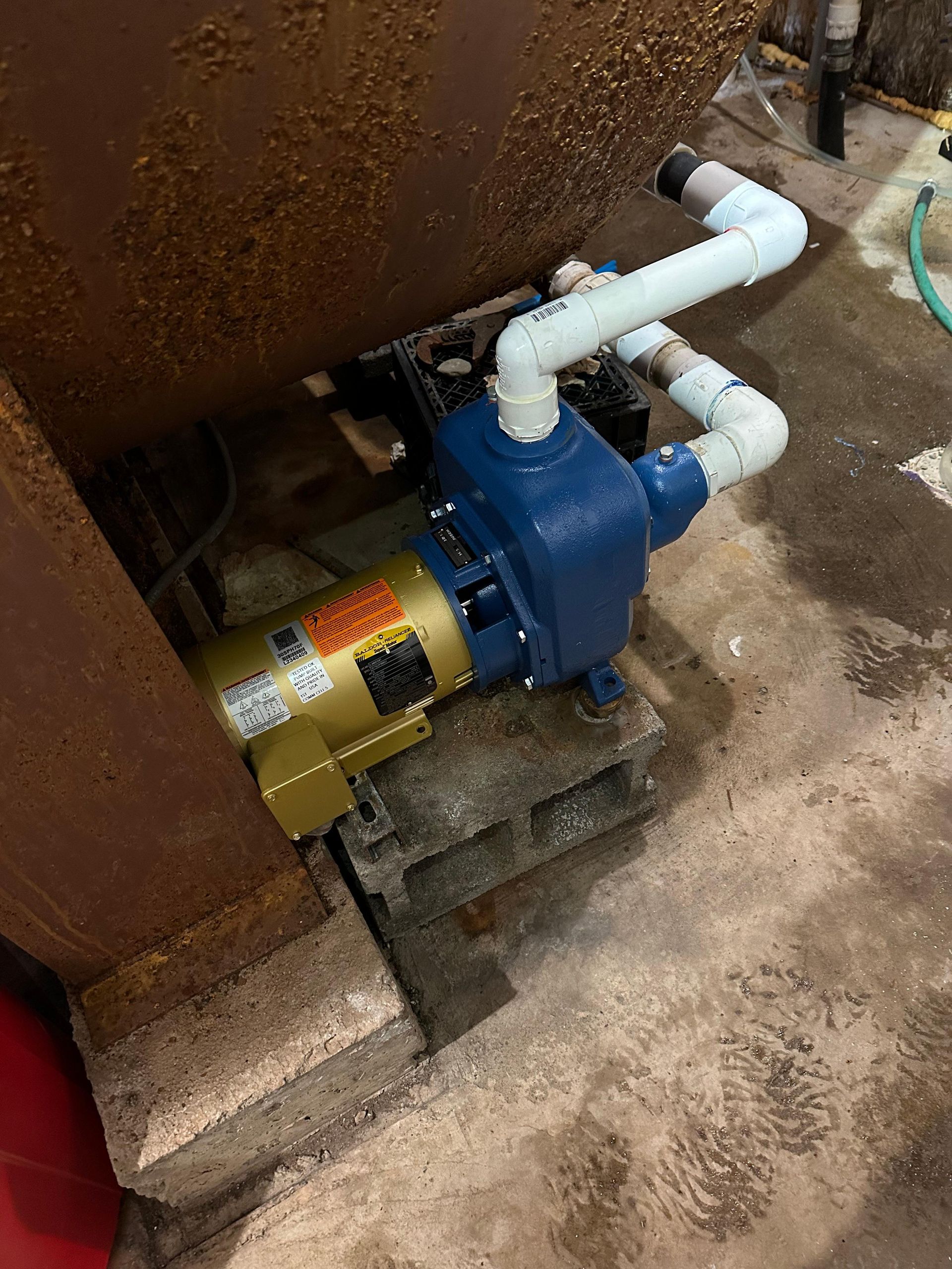 Blue and gold water pump on cinder blocks next to a rusty tank in a concrete-floored room.