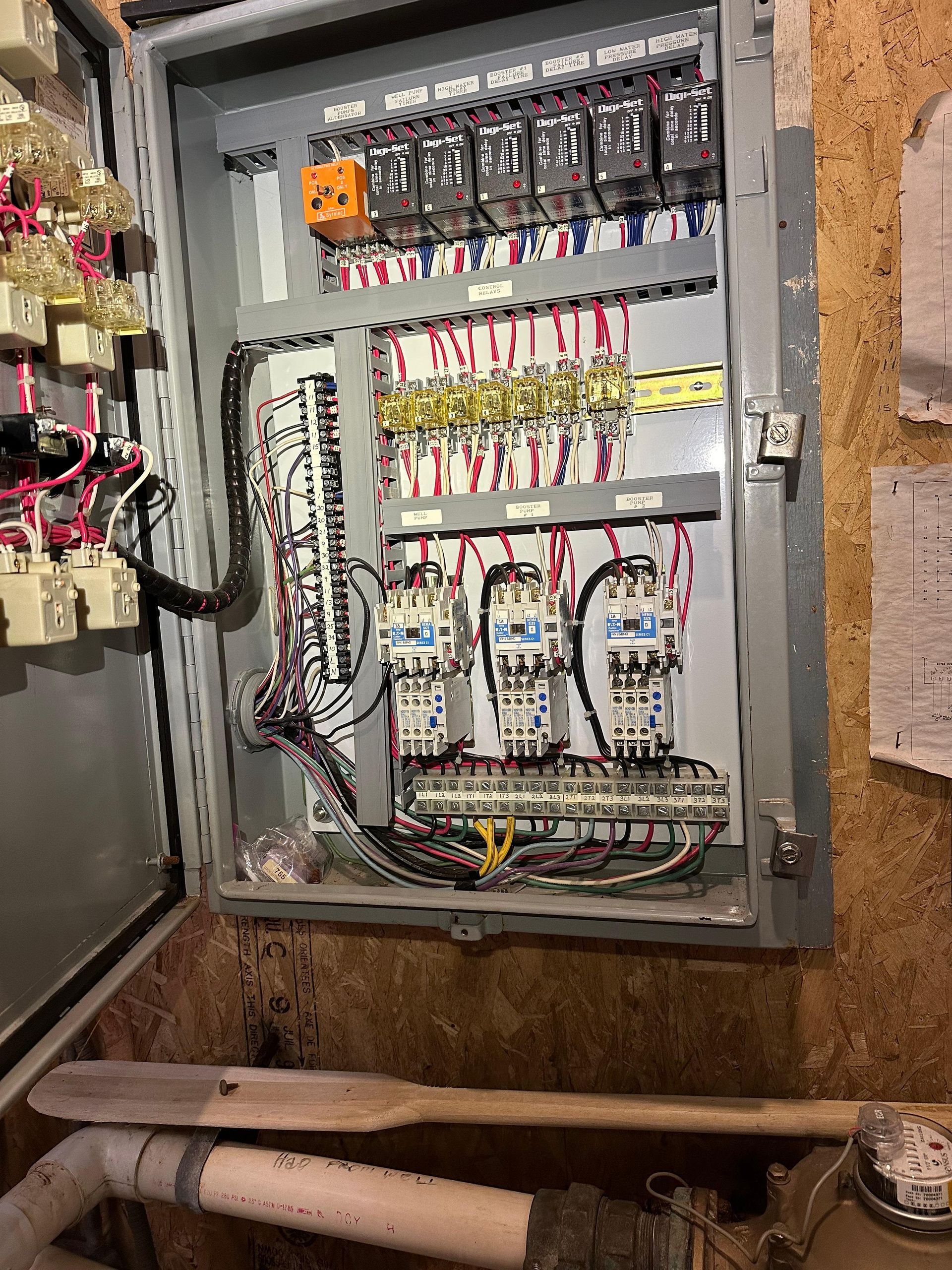 Open electrical control panel with various components and wiring mounted on a wall near plumbing.
