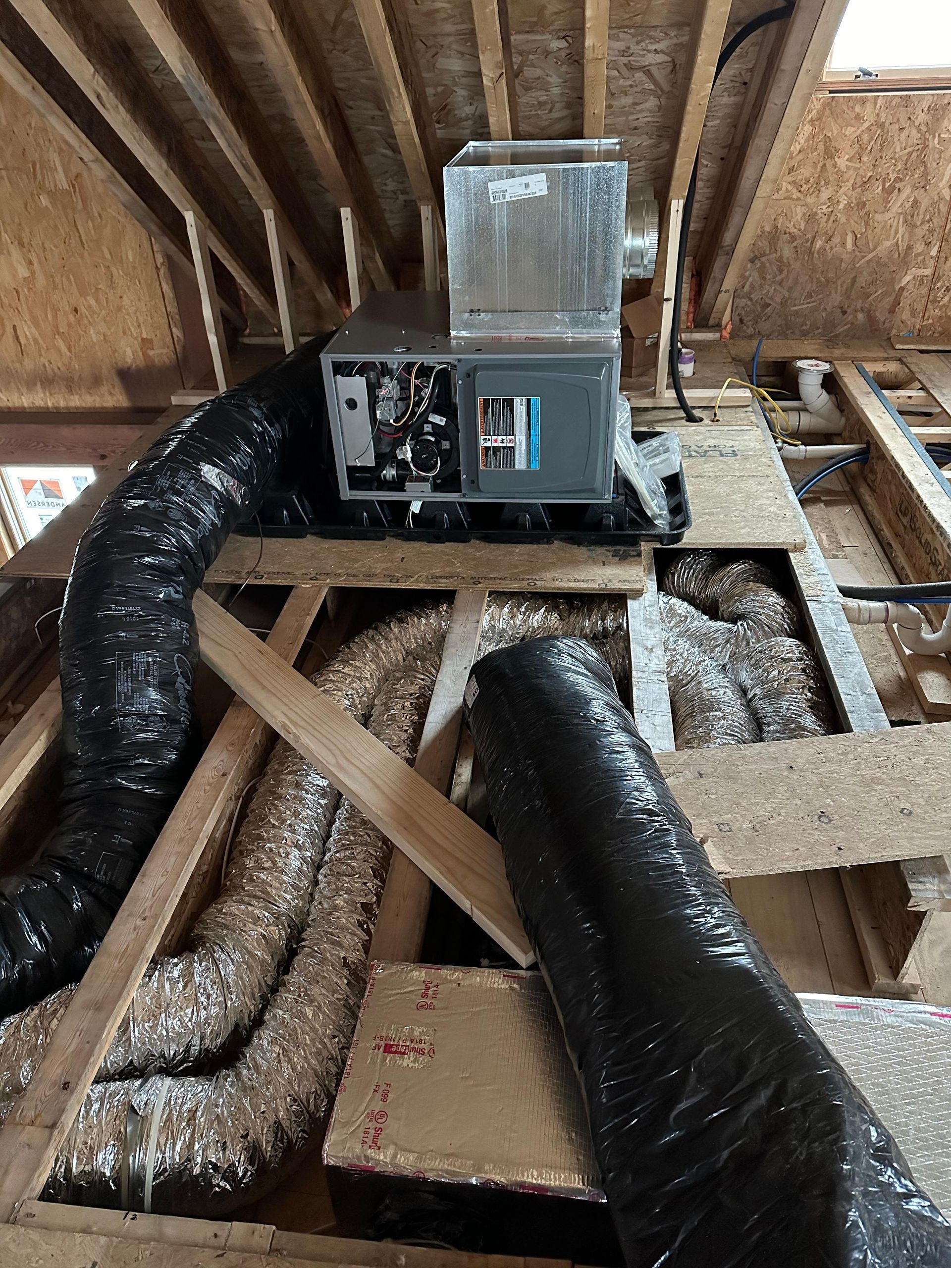 HVAC unit and ductwork installed in a wood-framed attic. Two black flexible ducts and one silver duct are visible.