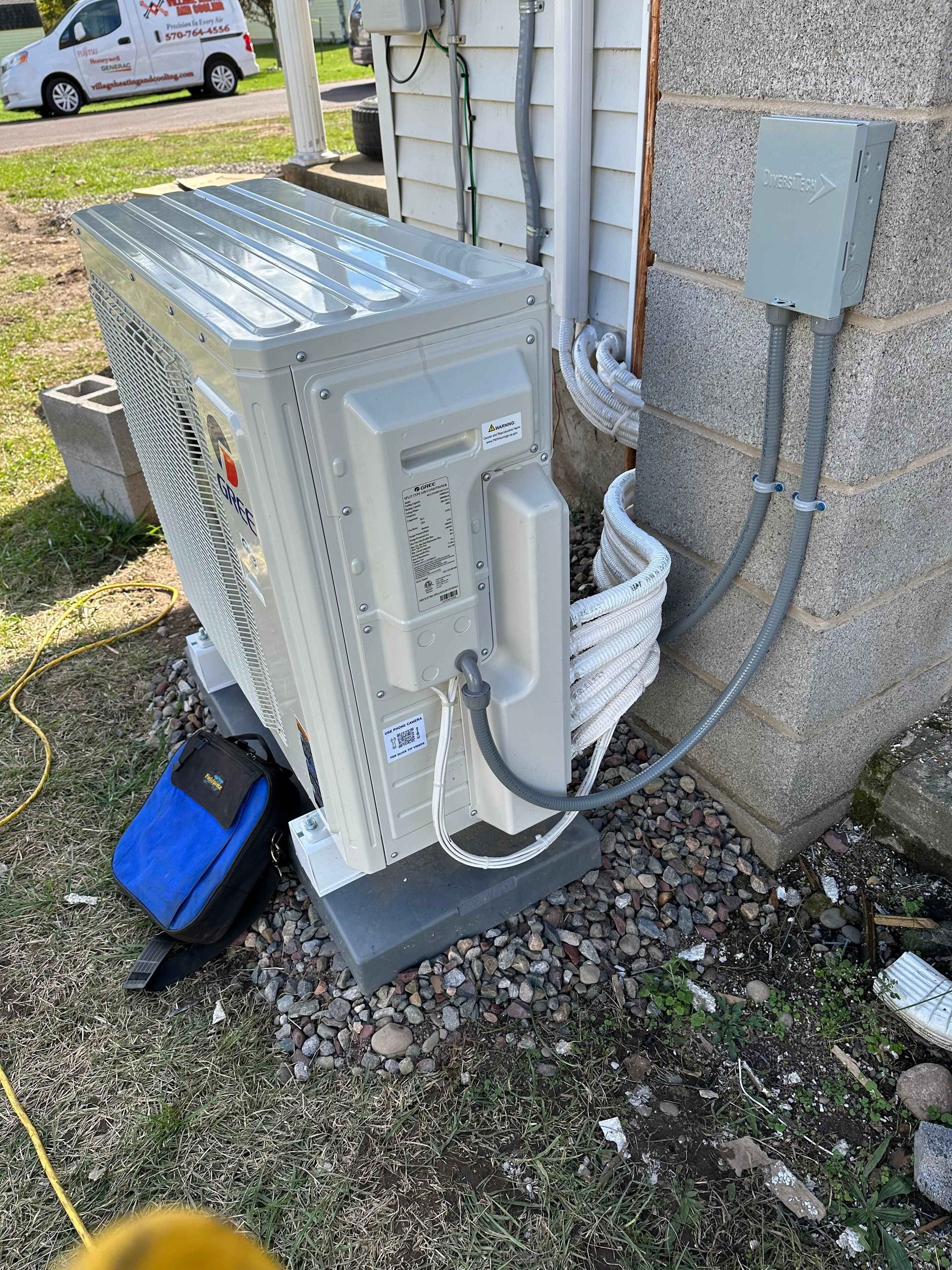 Exterior heat pump unit near a building; gray, white, and silver colors. Electrical box attached to the building.