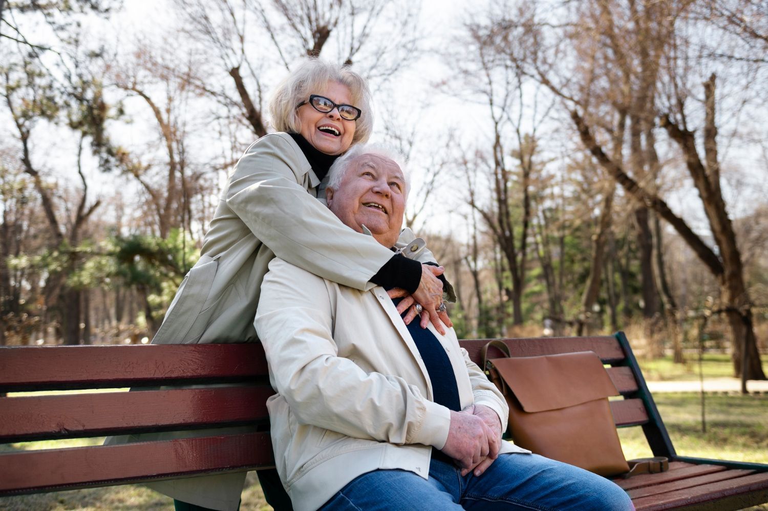 Two smiling people hugging on a park bench amid bare trees on a sunny day