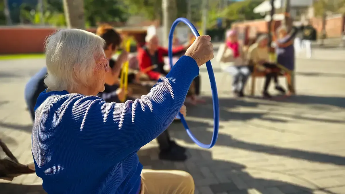 Mulher idosa com um suéter azul segura um arco azul em um parque, com outras pessoas próximas.