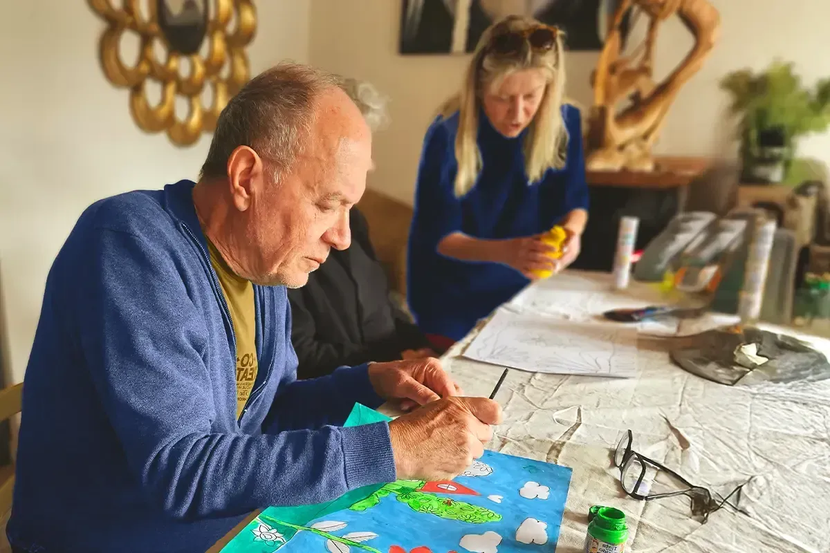 Homem pintando em uma mesa; mulher de blusa azul está atrás dele, segurando um recipiente amarelo.
