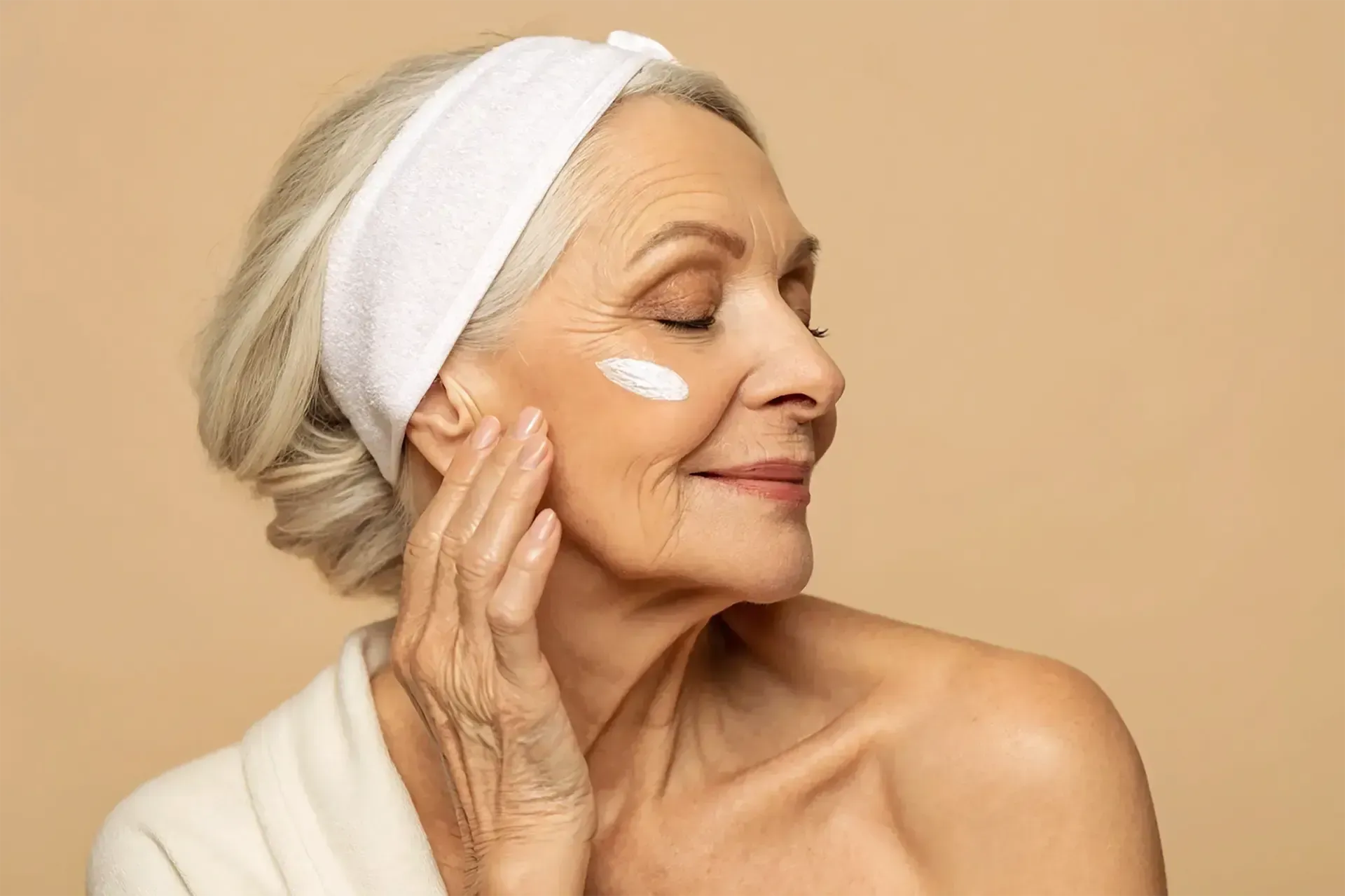Mulher de cabelos brancos aplicando creme no rosto, olhos fechados, sorrindo e satisfeita.
