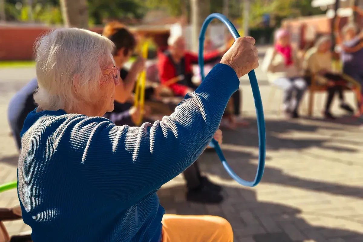 Grupo de pessoas em um lar de idosos se exercitando com bastões ao ar livre, sob a orientação de um instrutor.
