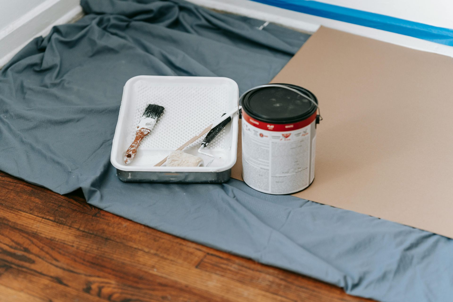 Painting supplies on a floor: tray with brushes, paint can, gray cloth, and beige board.