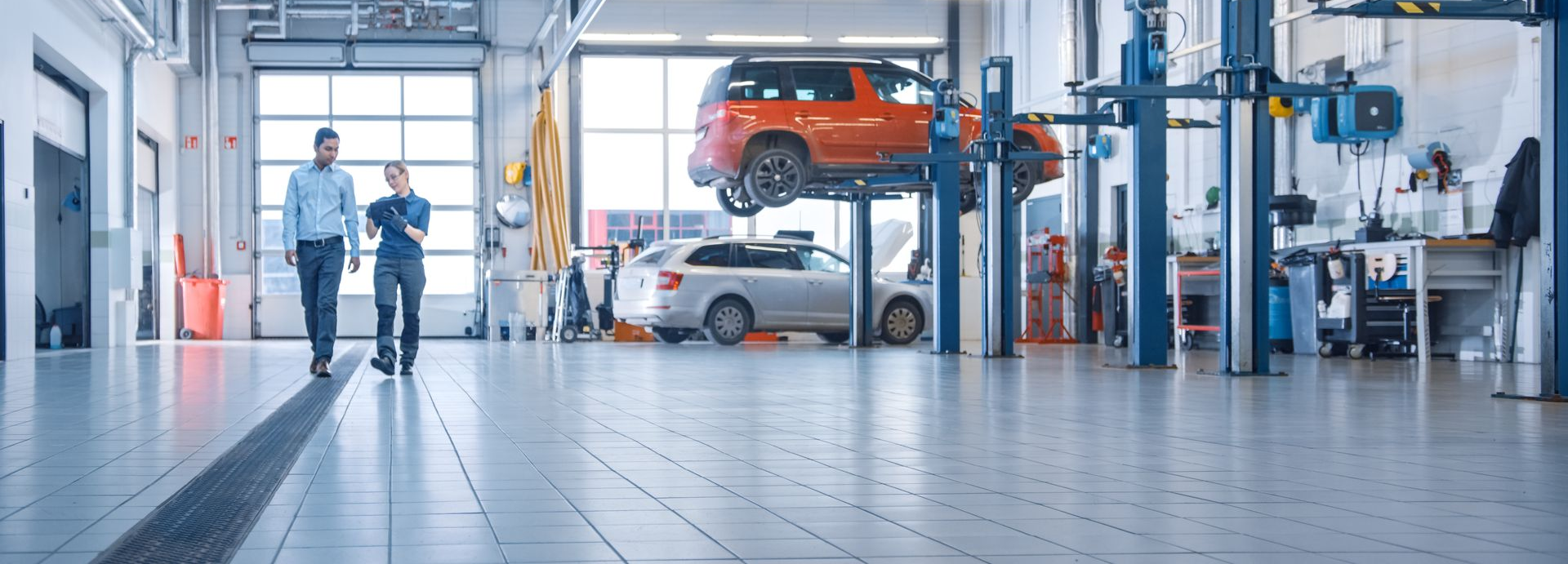 Two men walking in a bright auto repair shop; an orange SUV is lifted on a hoist.